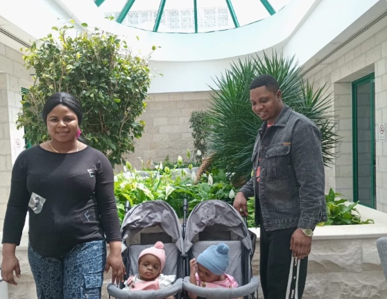 A woman and a man with two babies in strollers inside a bright indoor space with a glass ceiling, plants, and a stone wall.