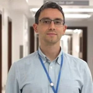 A man with glasses in a light blue button-up shirt and lanyard standing in a hallway with white walls and doors.