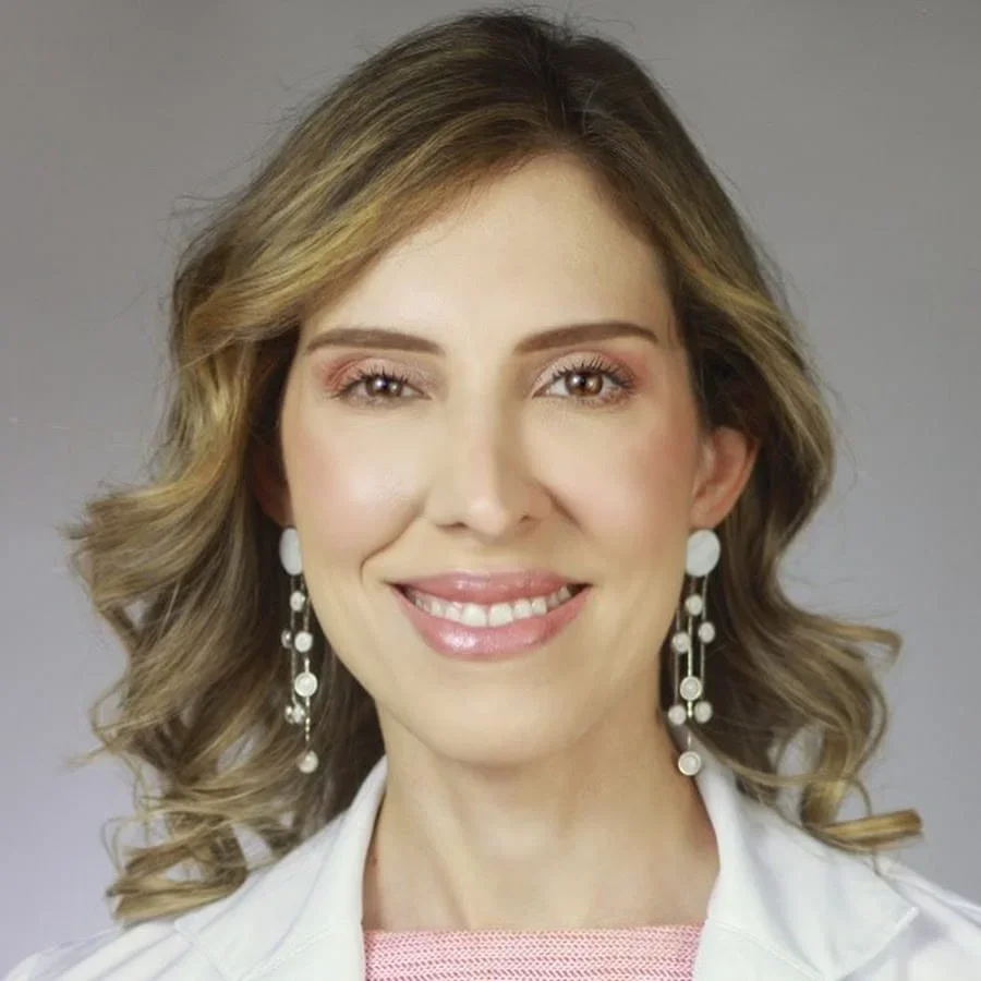 A woman with wavy blonde hair and makeup, wearing white pearl earrings and a white jacket, smiling at the camera against a plain background.