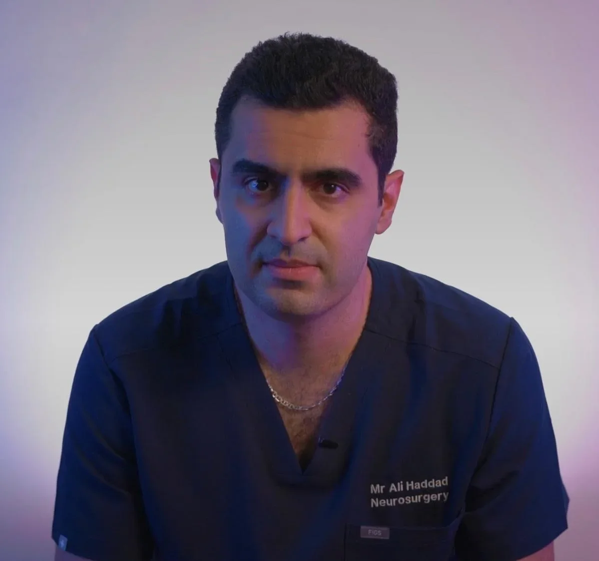 A man with dark hair and a serious expression, wearing a navy medical scrub top with and a gold chain, against a neutral background with purple and blue lighting.