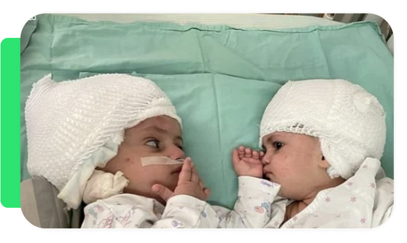 Two young children in hospital beds wearing white hats, lying face to face, touching hands, one with a nasal tube, in a hospital setting.