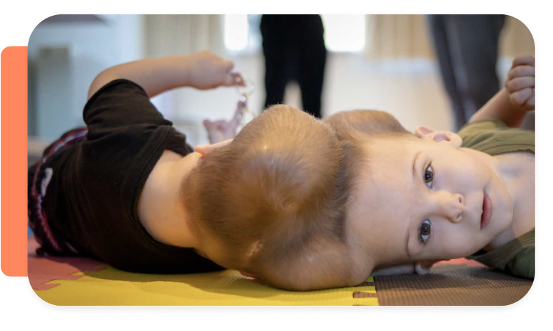 Child lying on the floor during a medical or therapy session with another person in the background.