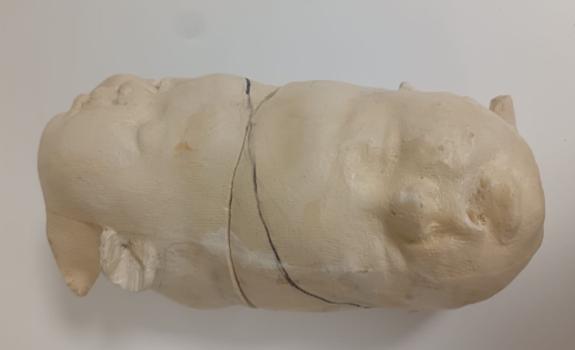 A large beige plastic or wax sculpture of a human head, oriented upside down with the top of the head at the bottom of the image, showing facial details such as closed eyes, nose, and lips.