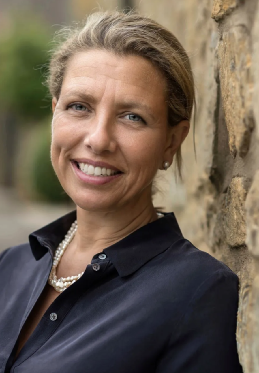 A woman with blonde hair styled back, wearing pearl earrings, a pearl necklace, and a black button-up shirt, smiling and leaning against a stone wall outdoors.