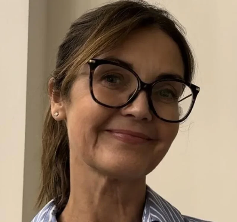 Close-up of a woman with short brown hair, wearing large black glasses, a small earring, and a light blue striped shirt, smiling softly.