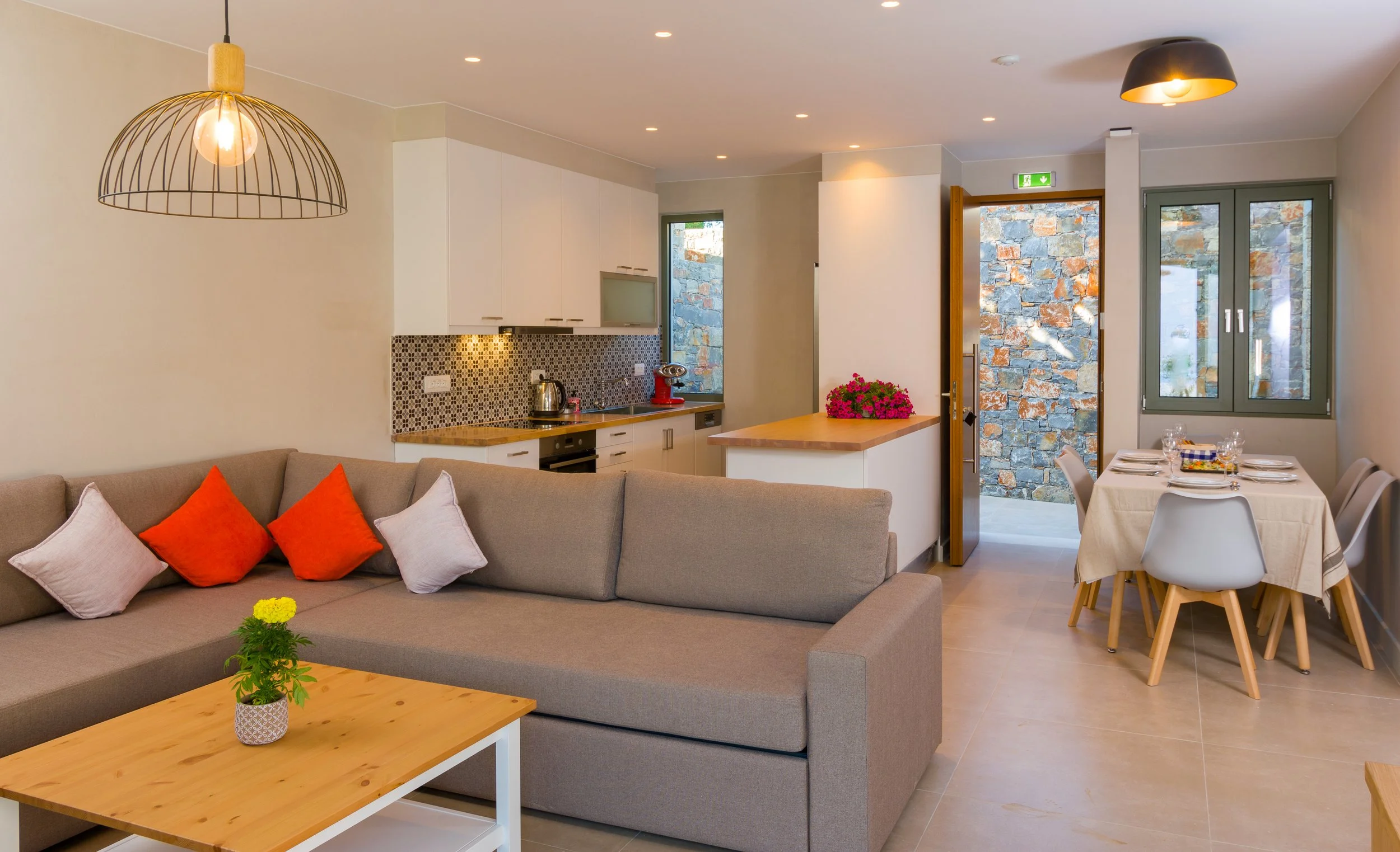 Living room and kitchen area with beige sofa with orange and white cushions, wooden coffee table with a small yellow flower, modern kitchen with white cabinets, colorful mosaic backsplash, dining table with white chairs, stone wall visible through op