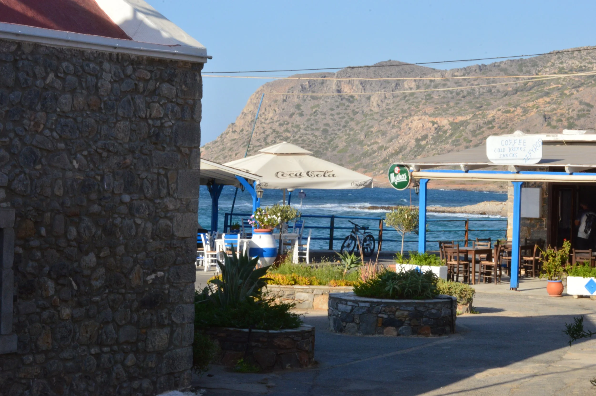 A seaside outdoor cafe area with tables, chairs, umbrellas, and potted plants. In the background, a rocky mountain and clear blue sky are visible. A stone wall partially blocks the view on the left side.