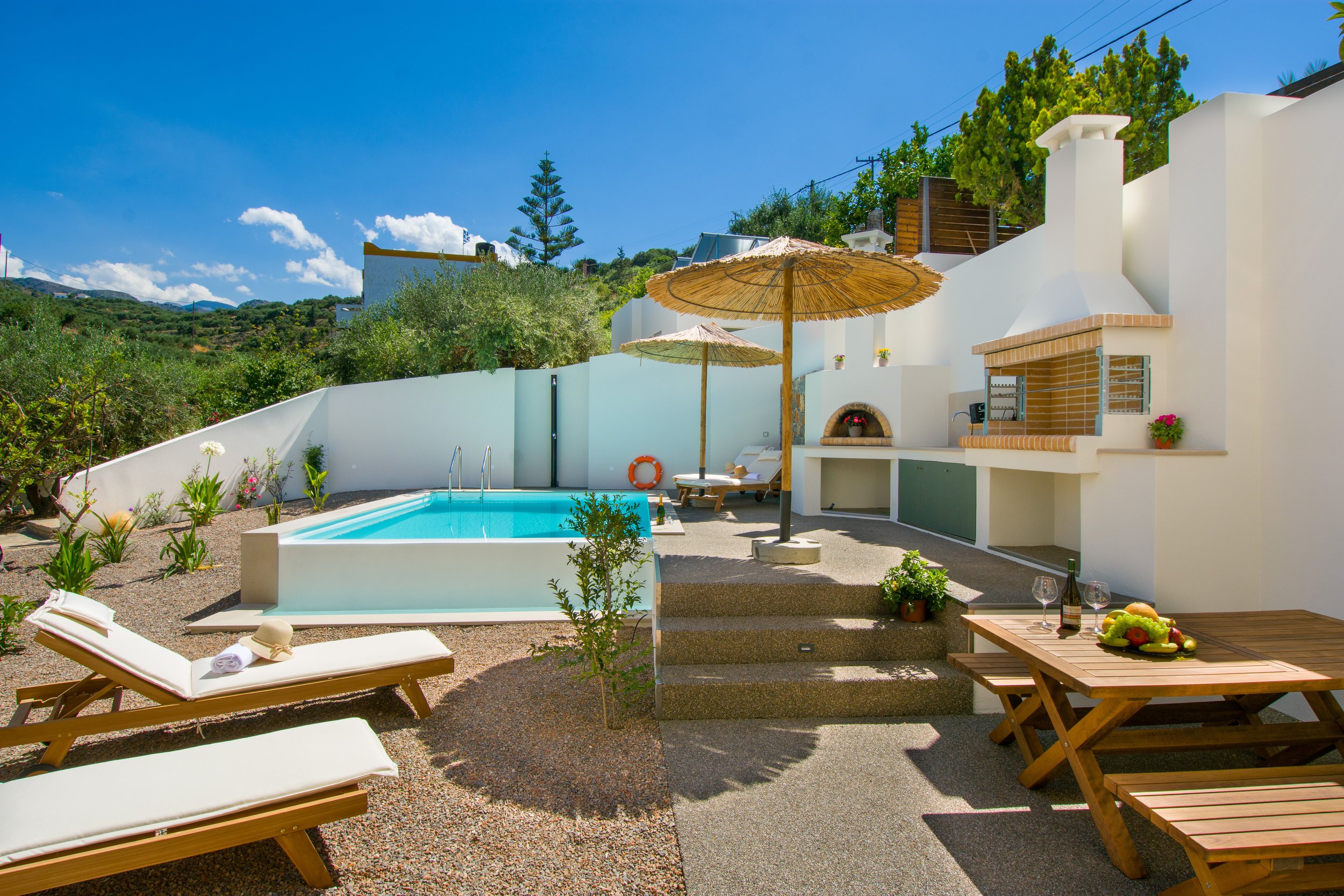A sunny outdoor pool area with lounge chairs, umbrellas, and a view of lush green hills and trees against a bright blue sky.