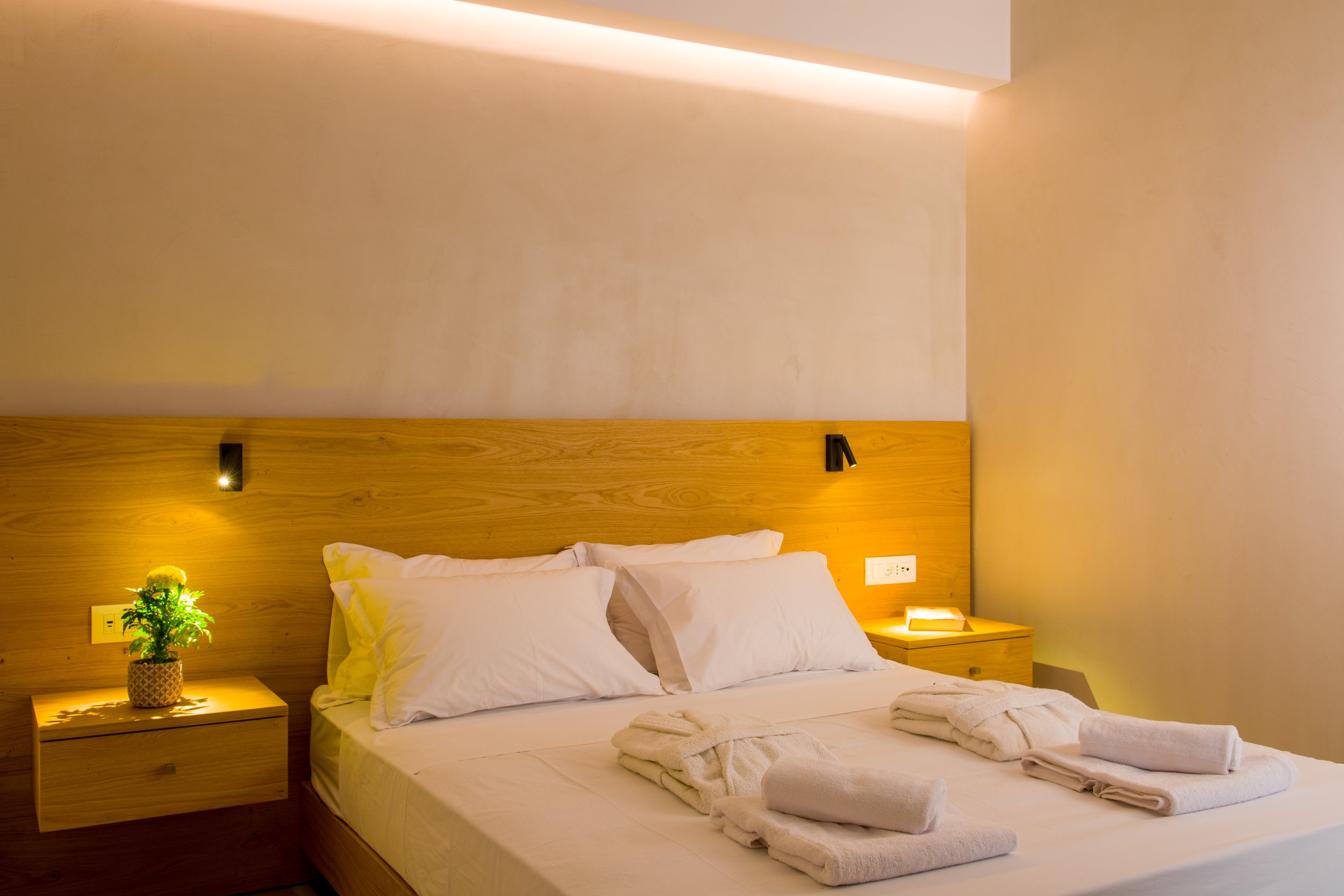 A neatly made modern hotel bed with white linens and two pillows, flanked by wooden nightstands with lamps and green plants, against a beige wall with subtle lighting