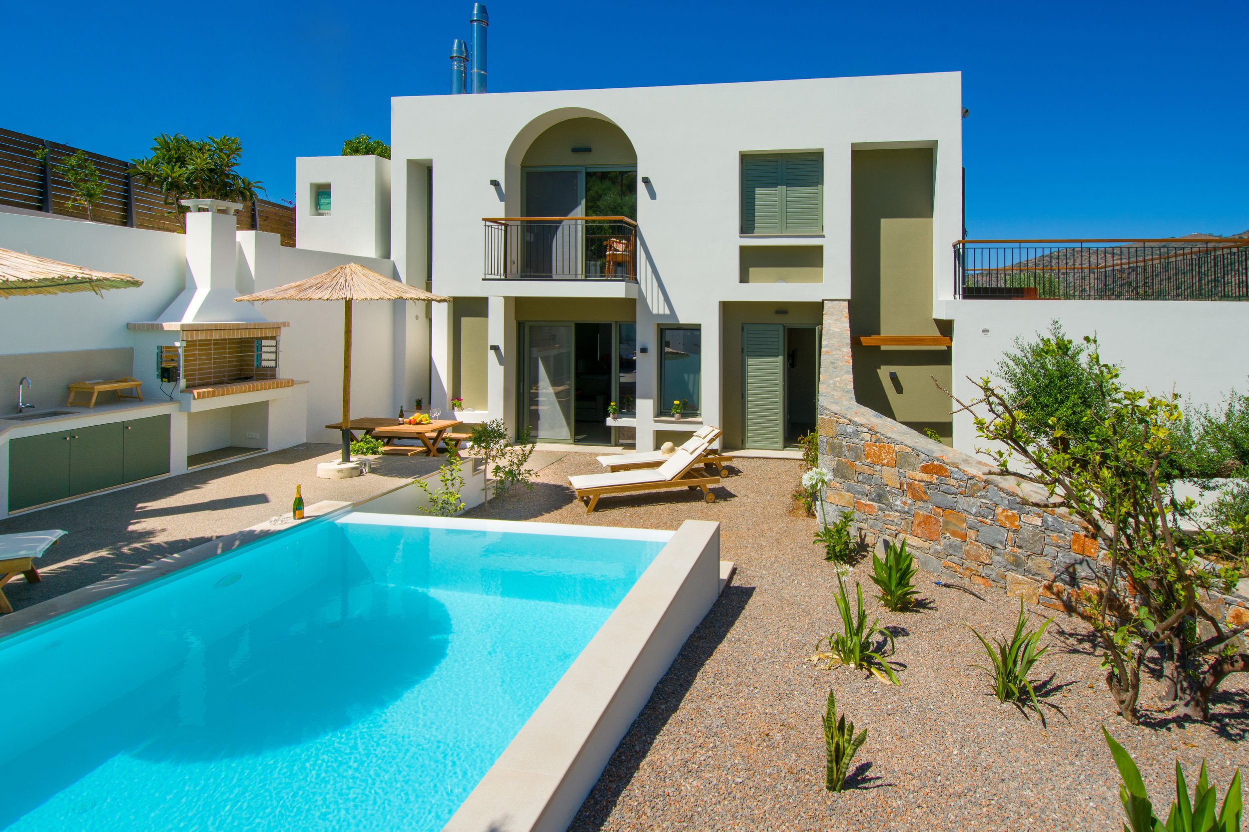 Milatos Garden Villas - Modern white house with a backyard pool, lounge chairs, umbrellas, and outdoor kitchen area under clear blue sky.