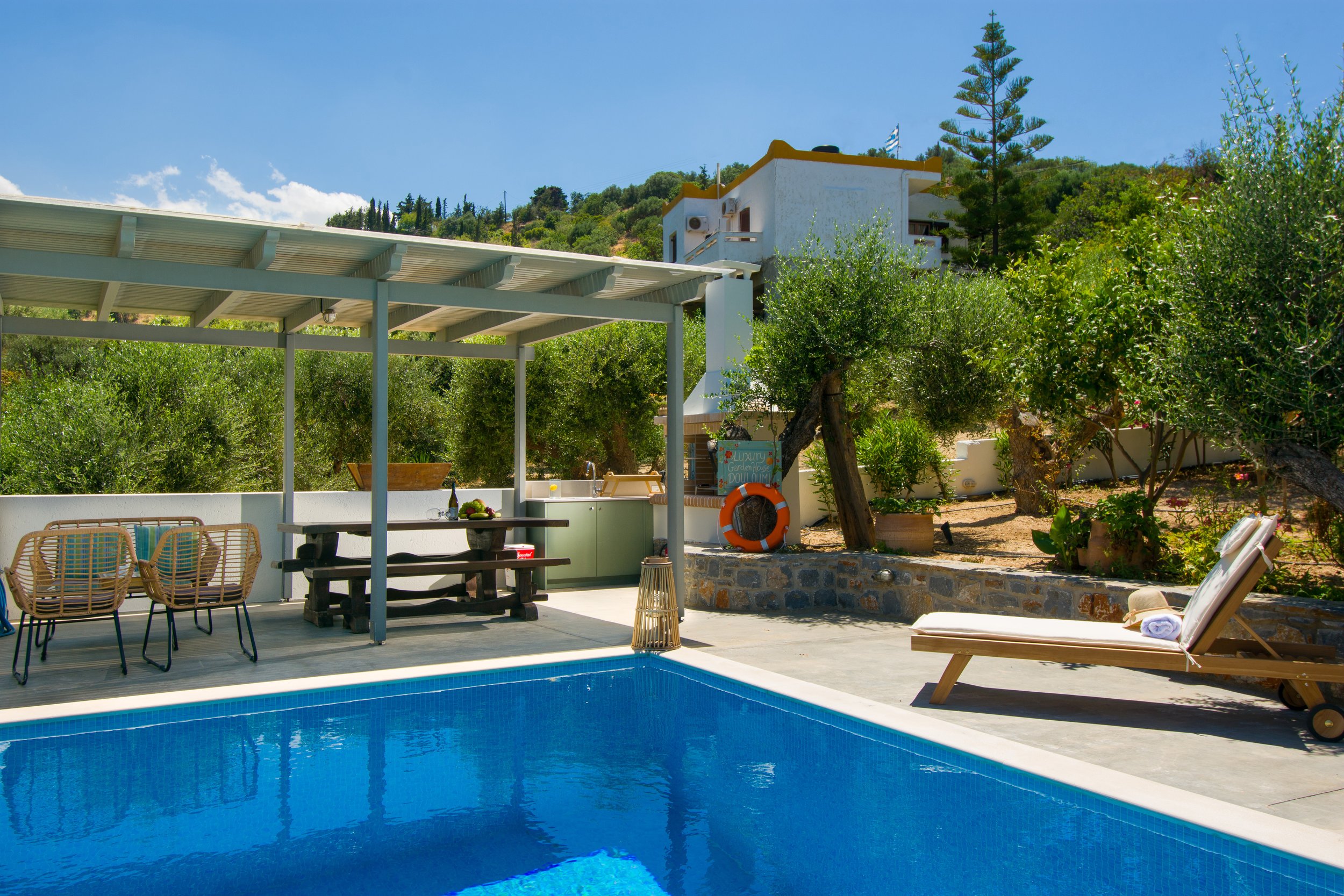 A backyard with a swimming pool, outdoor furniture, surrounding greenery, and a white house on a hillside under a blue sky.