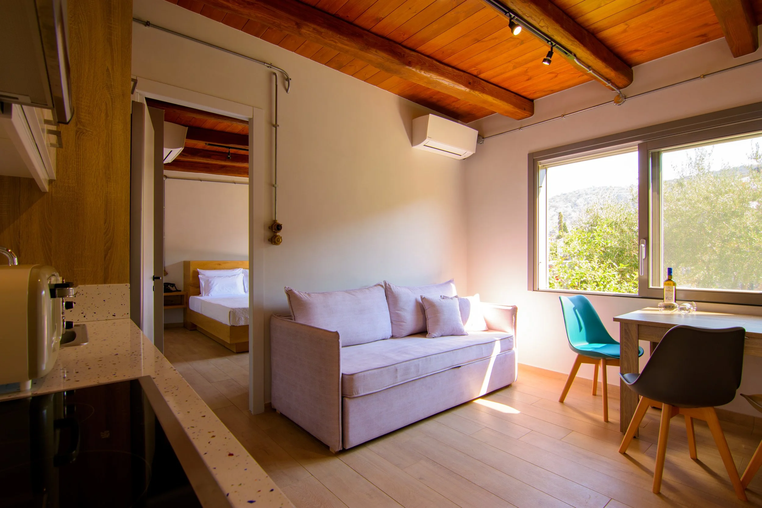 A cozy apartment interior with a wooden ceiling, large window showing greenery outside, a beige sofa, a wooden dining table with colorful chairs, a bed visible through a doorway, and a small kitchen area with appliances.