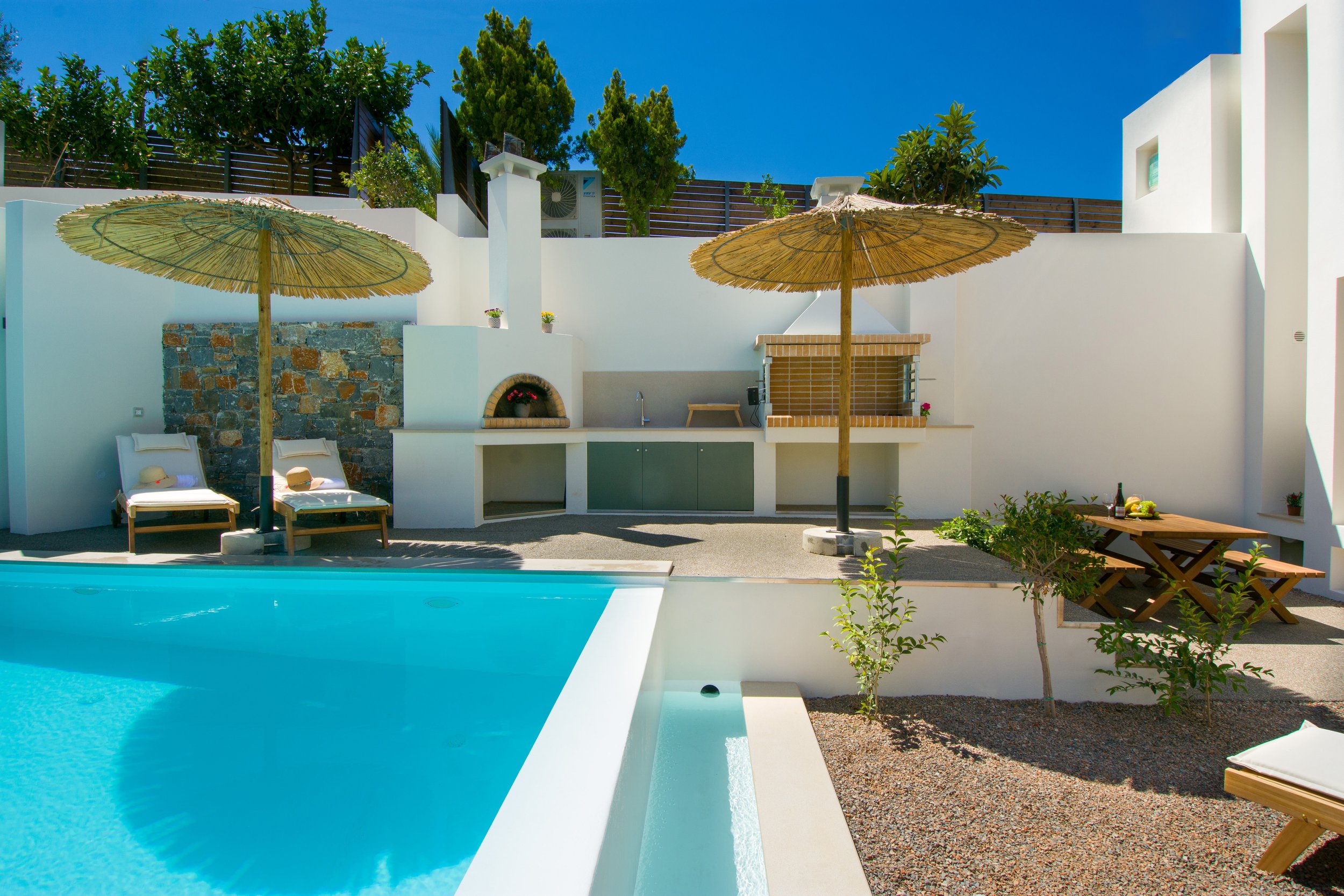 A bright outdoor pool area with two straw umbrellas, white lounge chairs with hats, a built-in barbecue, and lush greenery against a white modern wall and blue sky.