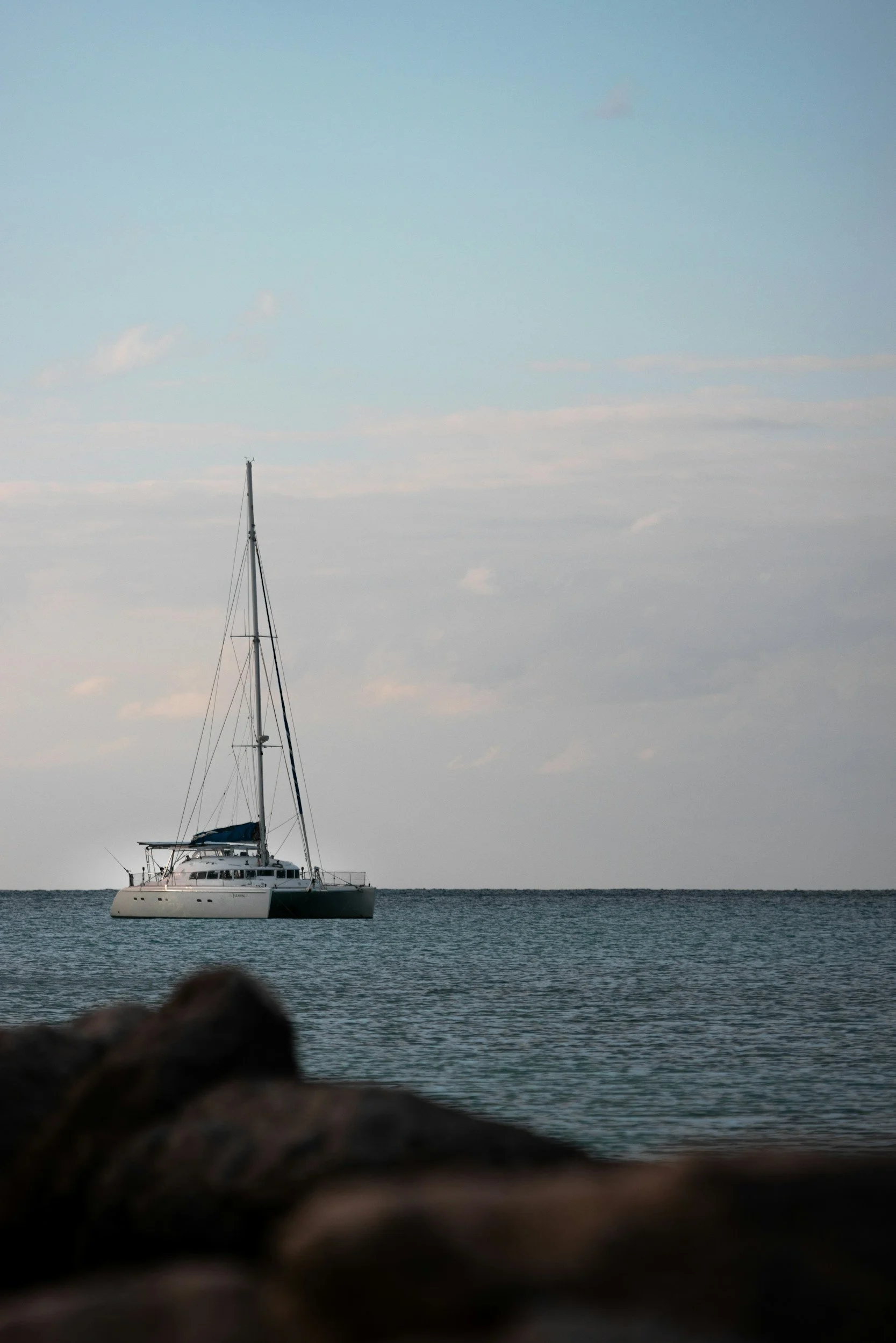A sailboat with a dark hull and tall mast floats on calm water under a sky with soft clouds.