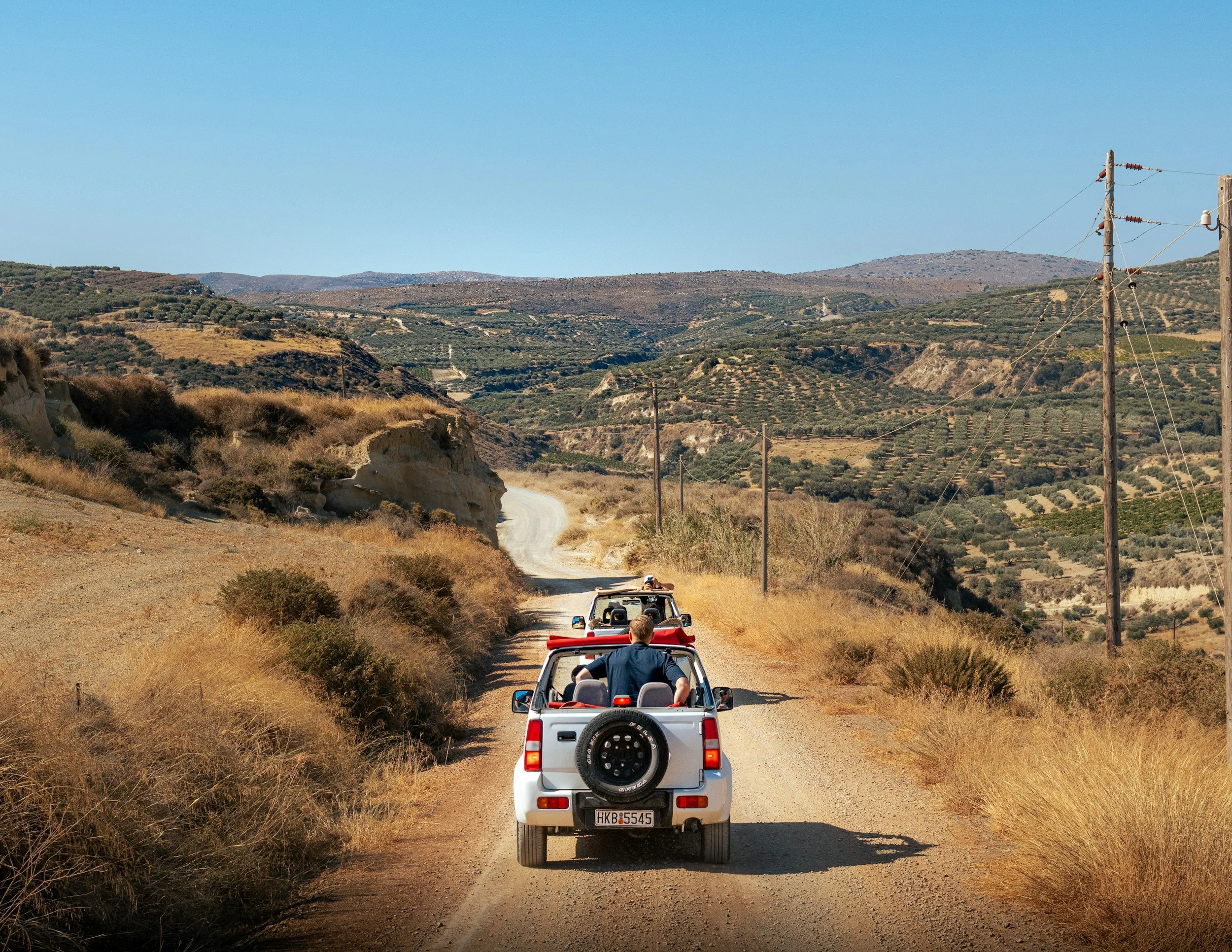 A convoy of white off-road vehicles driving on a dirt road through a hilly, semi-arid landscape with power lines on the right side of the road and dry grass on both sides.