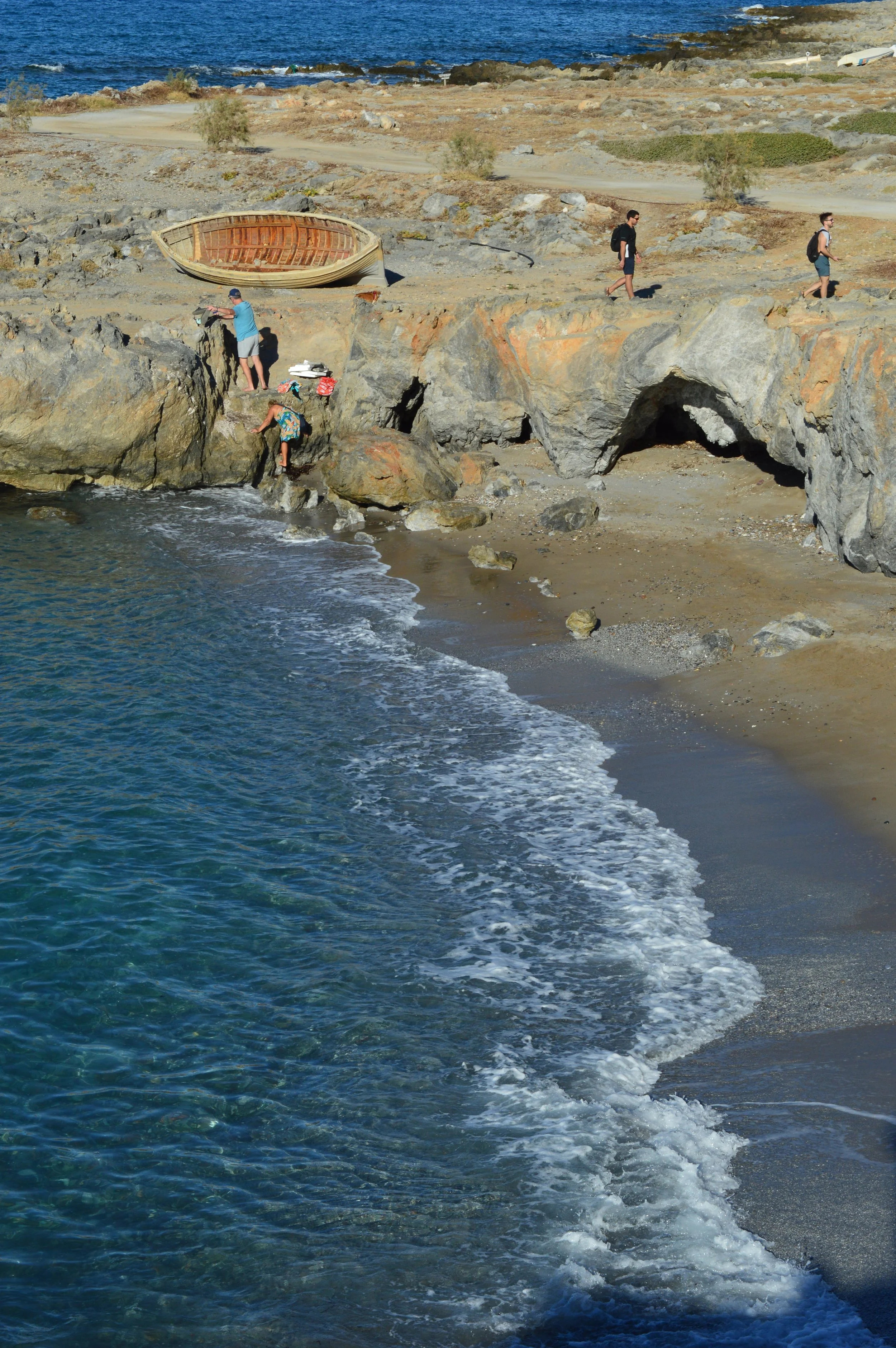 Milatos Garden Villas -  Aspa Beach - A coastal scene with rocky cliffs and a shoreline. A small boat is turned upside down on the rocks. Several people are walking along the rocky shore, with some climbing rocks near the water.