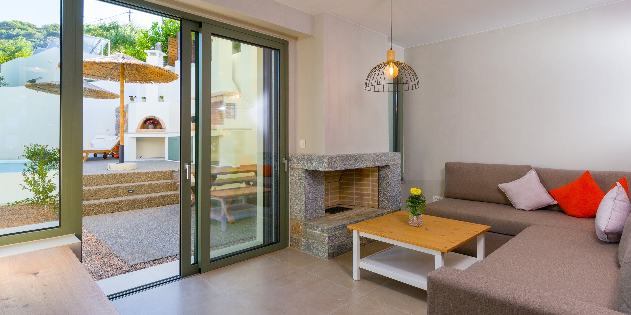 Living room with sliding glass door leading to outdoor patio with umbrellas, lounge chairs, and plants. Interior features a gray sofa with colorful pillows, a wooden coffee table with a potted plant, a fireplace, and a hanging light fixture.