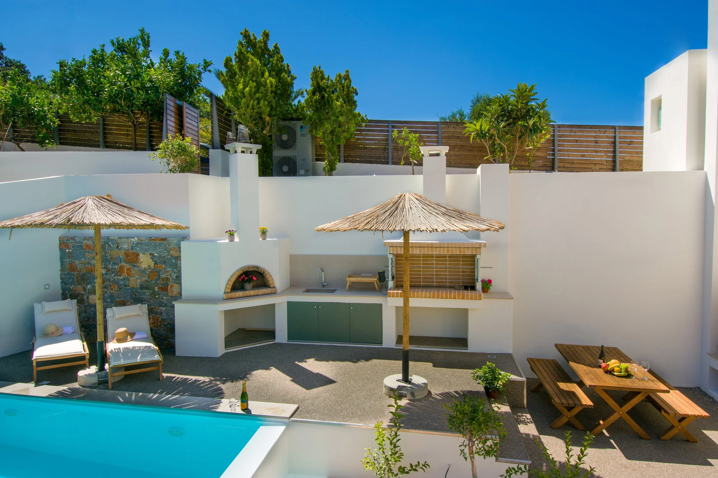 Views of a backyard with a pool, white wall, thatched umbrellas, lounge chairs, and a wooden table with fruit and drinks. Green trees and a wooden fence are in the background under a clear blue sky.