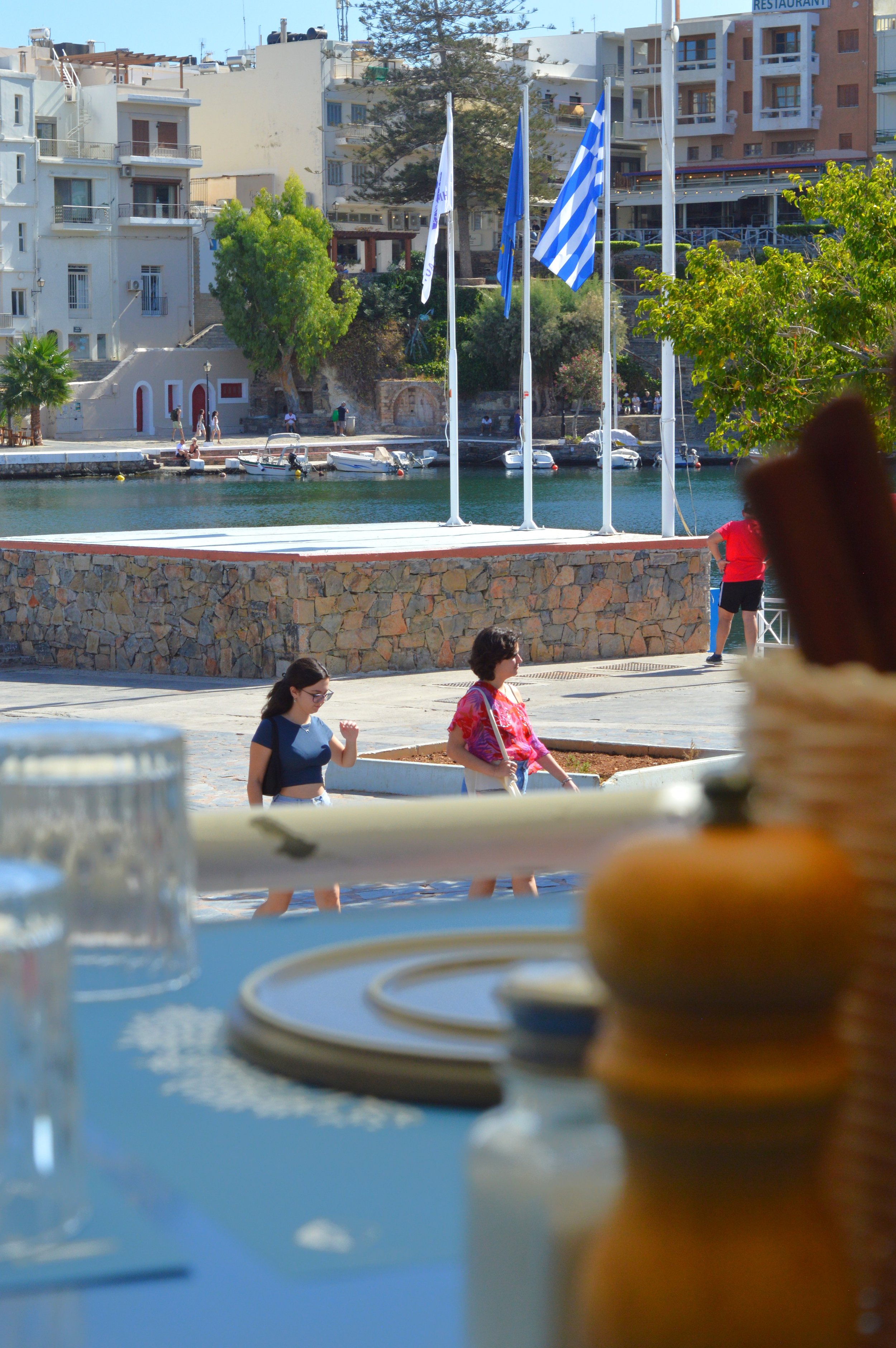 Milatos Garden Villas - Karnagio rest - A waterfront scene with boats docked along the water, multi-story buildings in the background, and people walking along the waterfront path. There are prominent blue and white flags flying in the middle ground.