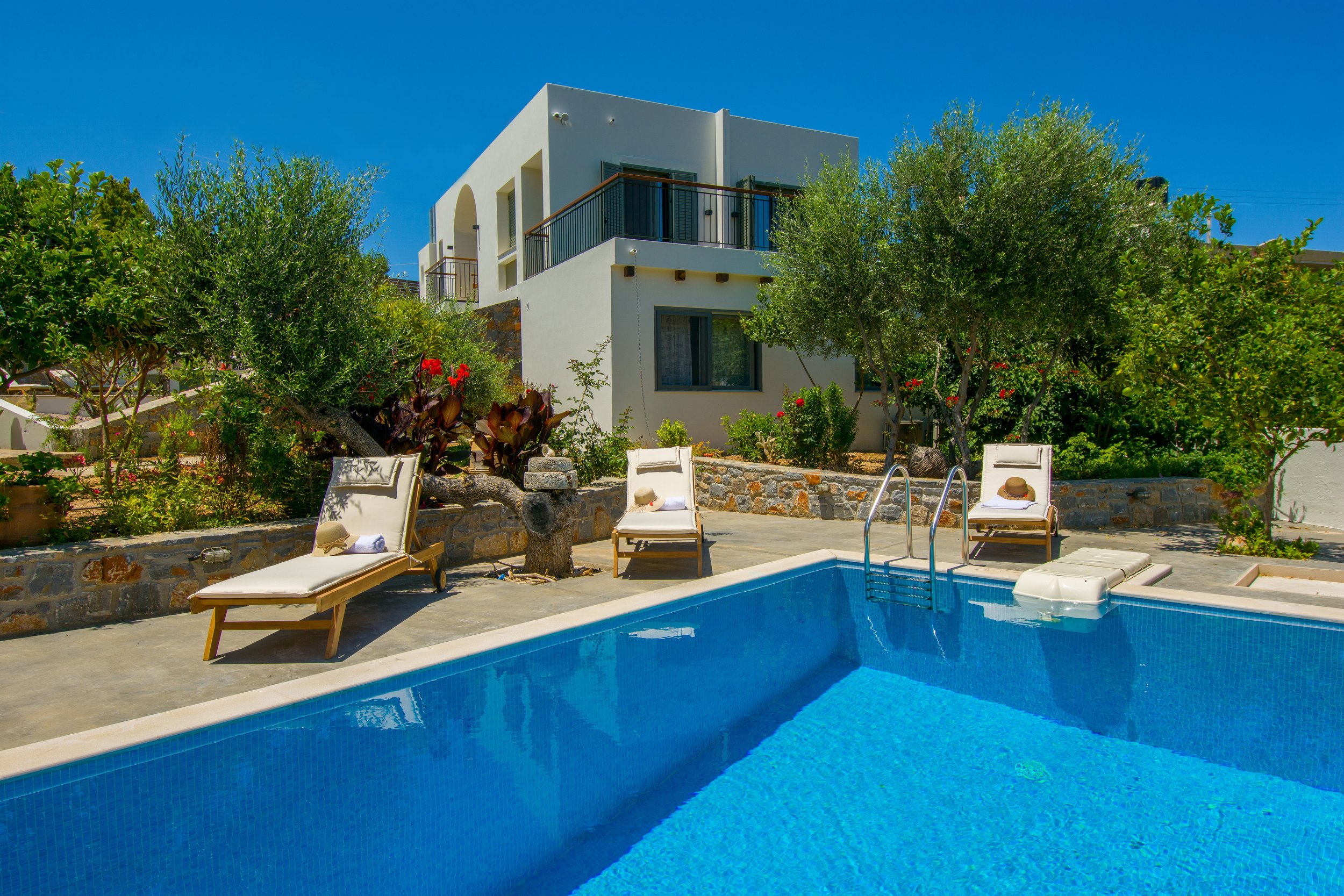 A modern white house with balconies, surrounded by lush green trees and red flowers, with a swimming pool in the foreground. There are sun loungers with hats and towels near the pool.
