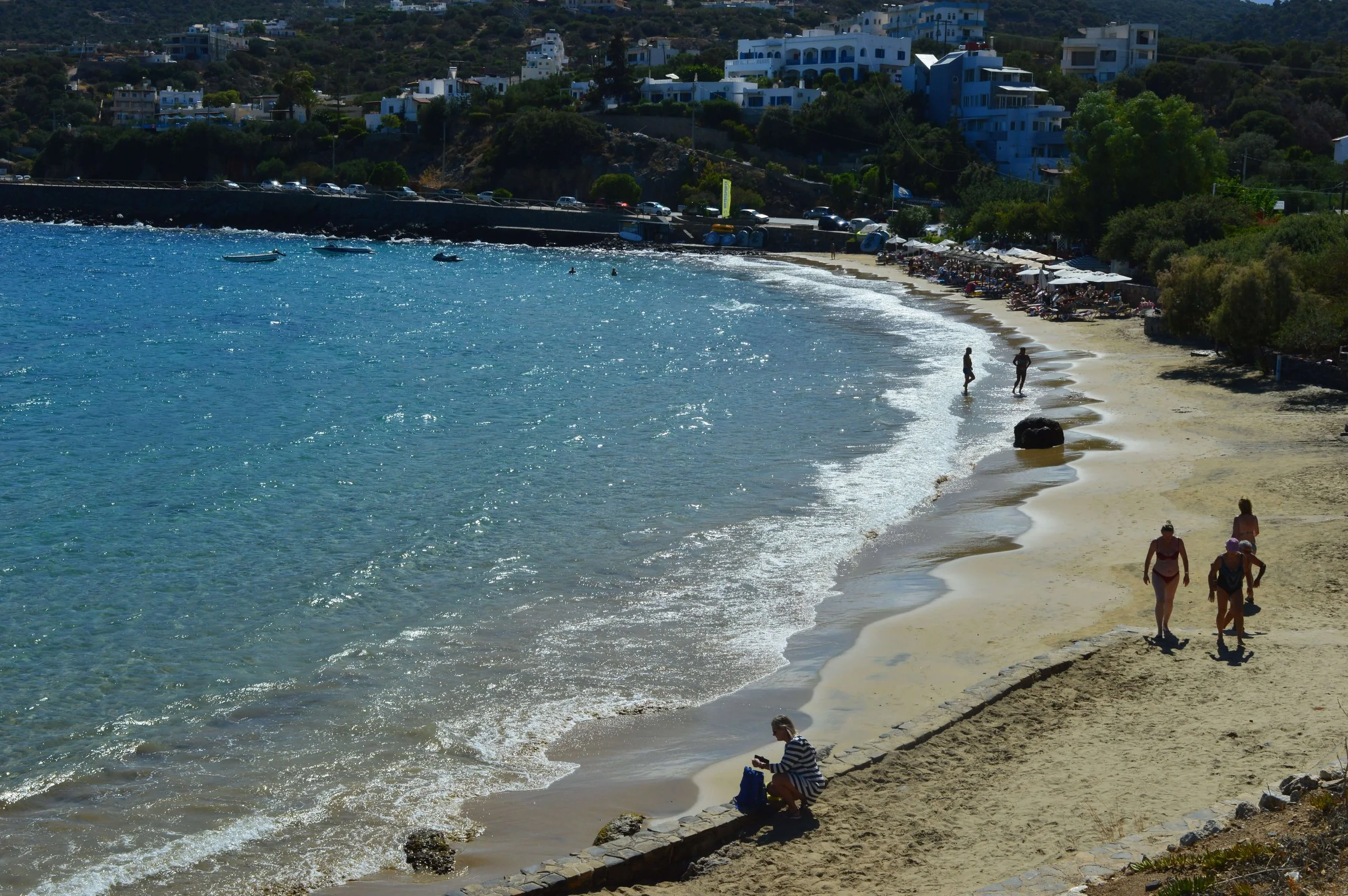 People walking and sitting on a sandy beach with umbrellas, near turquoise water, with white buildings on a hillside in the background.