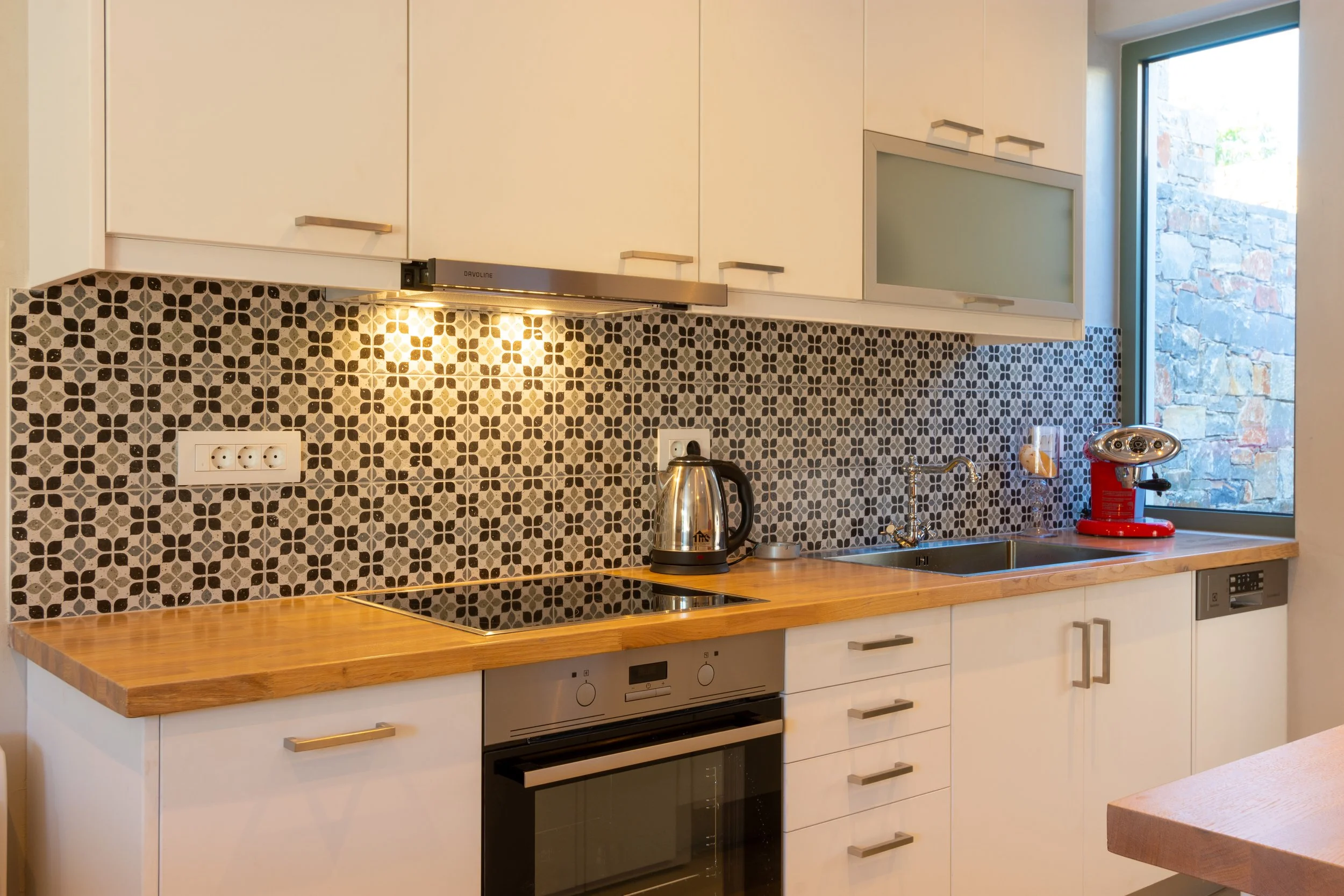 A modern kitchen with white cabinets, patterned black and white tile backsplash, wooden countertop, window showing stone wall outside, with kitchen appliances including an electric kettle, a red fire extinguisher, and a coffee maker.
