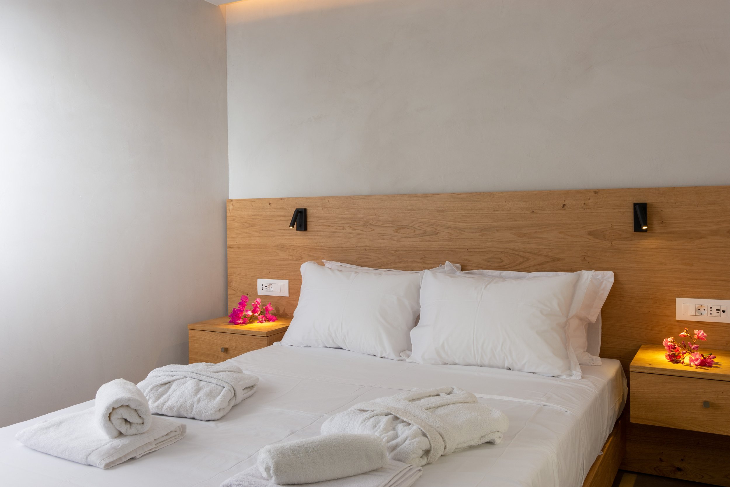 A neatly made bed with white linens and pillows, two rolled towels, pink flowers on both nightstands, modern black wall-mounted reading lights, and a wooden headboard in a minimalistic room.
