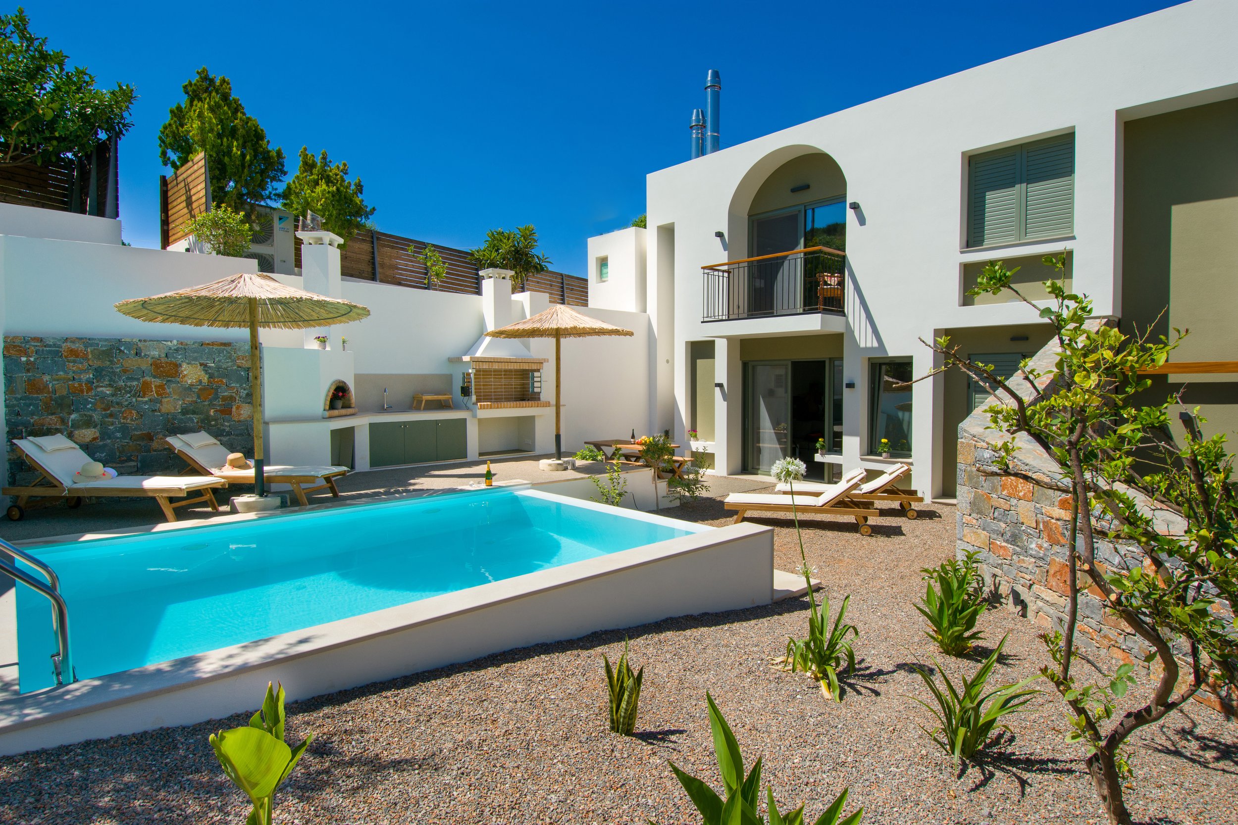 A modern house with white exterior walls featuring a swimming pool with blue water, lounge chairs with hats, and a patio area with umbrellas. The house has a balcony, green plants, and a clear blue sky.