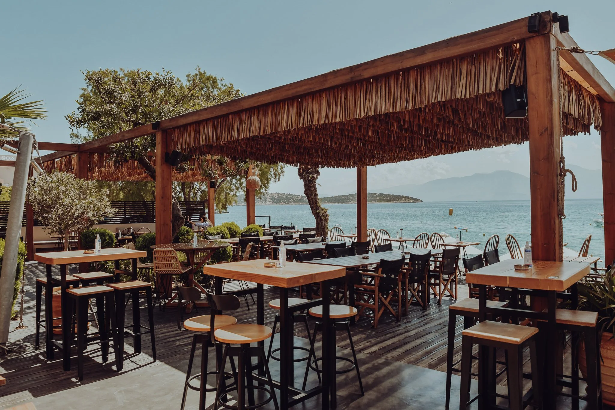 An outdoor restaurant with wooden tables, black chairs, and a thatched roof structure overlooking the water with trees and mountains in the background.