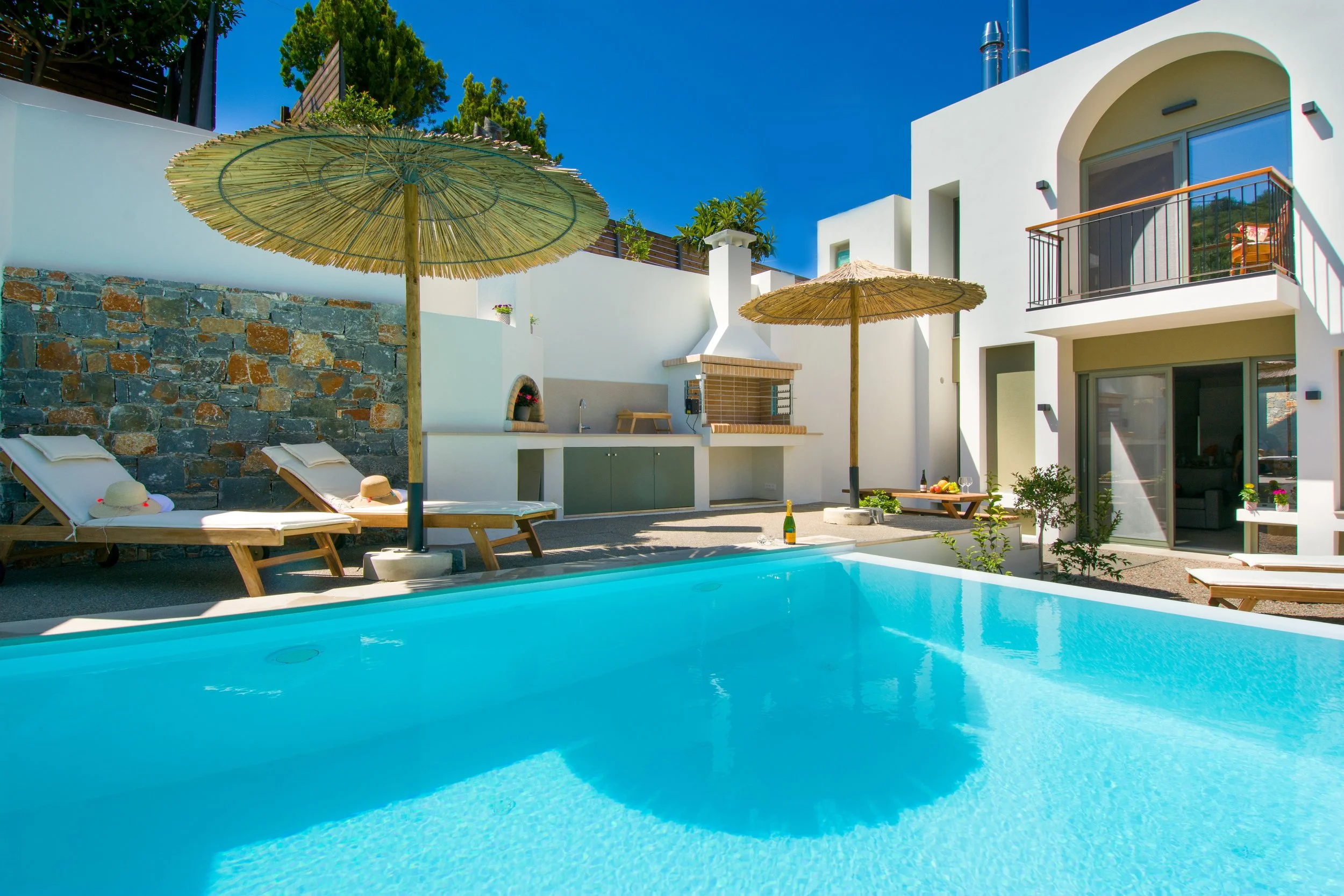 A swimming pool in a modern white house backyard with patio chairs, straw umbrellas, and an outdoor kitchen, under a clear blue sky.
