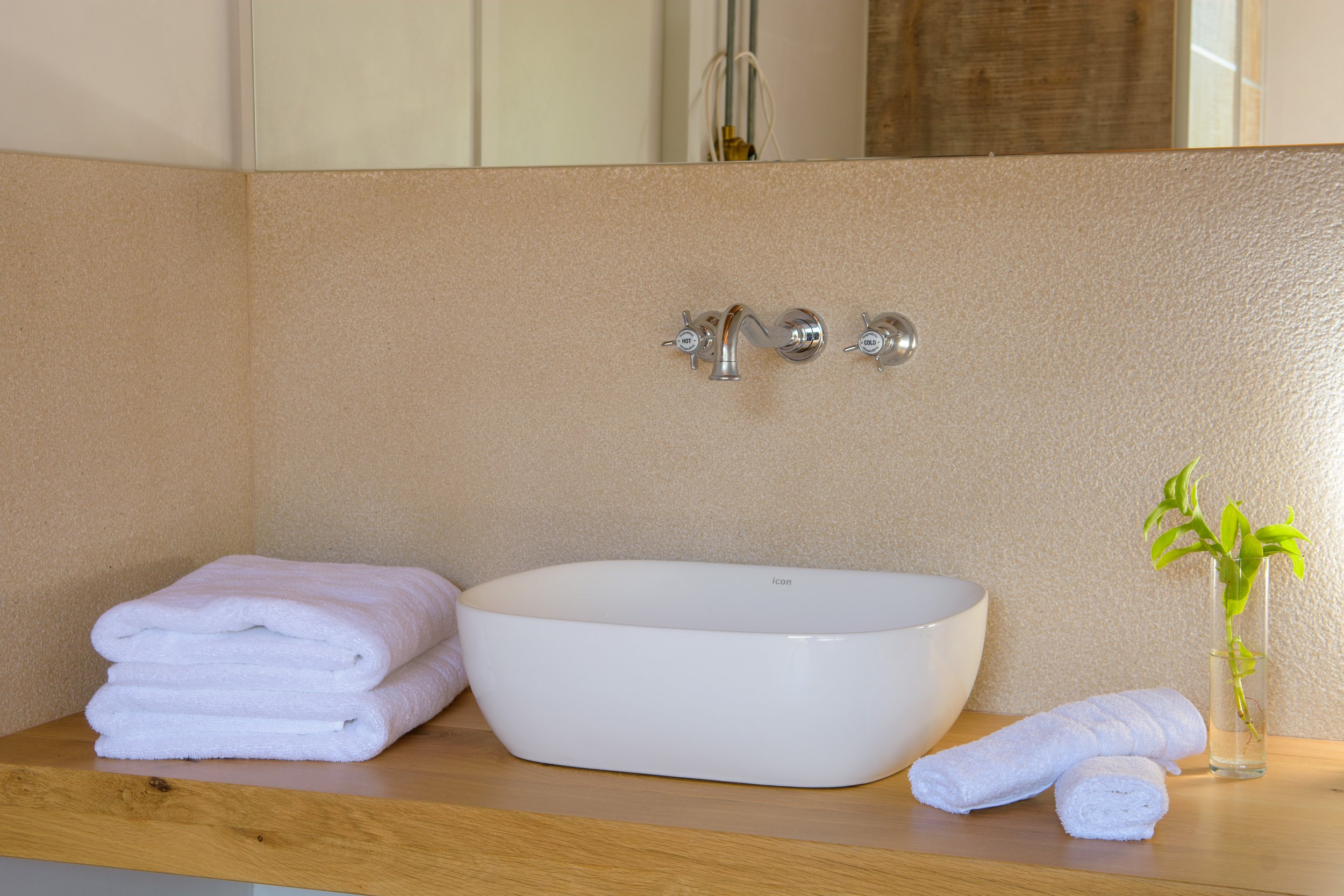 A bathroom decor setup featuring a white ceramic sink on a wooden countertop, with neatly folded white towels beside it, a small potted green plant on the right, and a mirror and textured beige wall above.