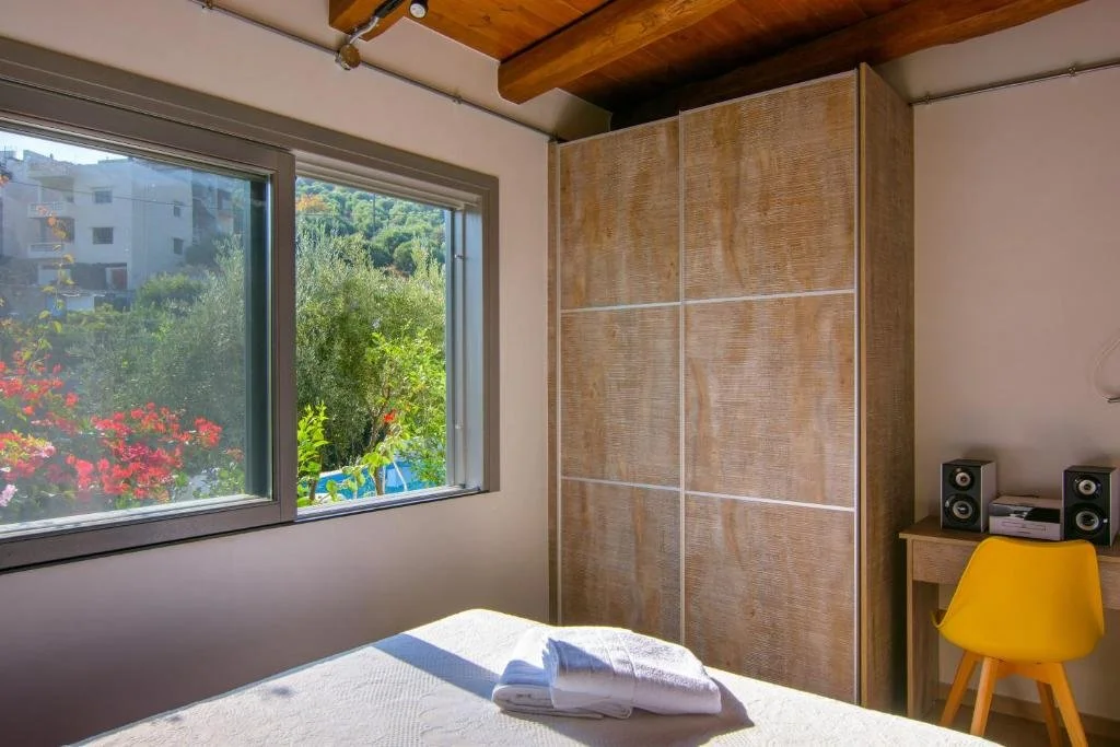 A bedroom with a large window showing a garden with greenery and flowers, a bed with folded towels, a wardrobe with sliding doors, a wooden desk with two speakers, and a yellow chair.