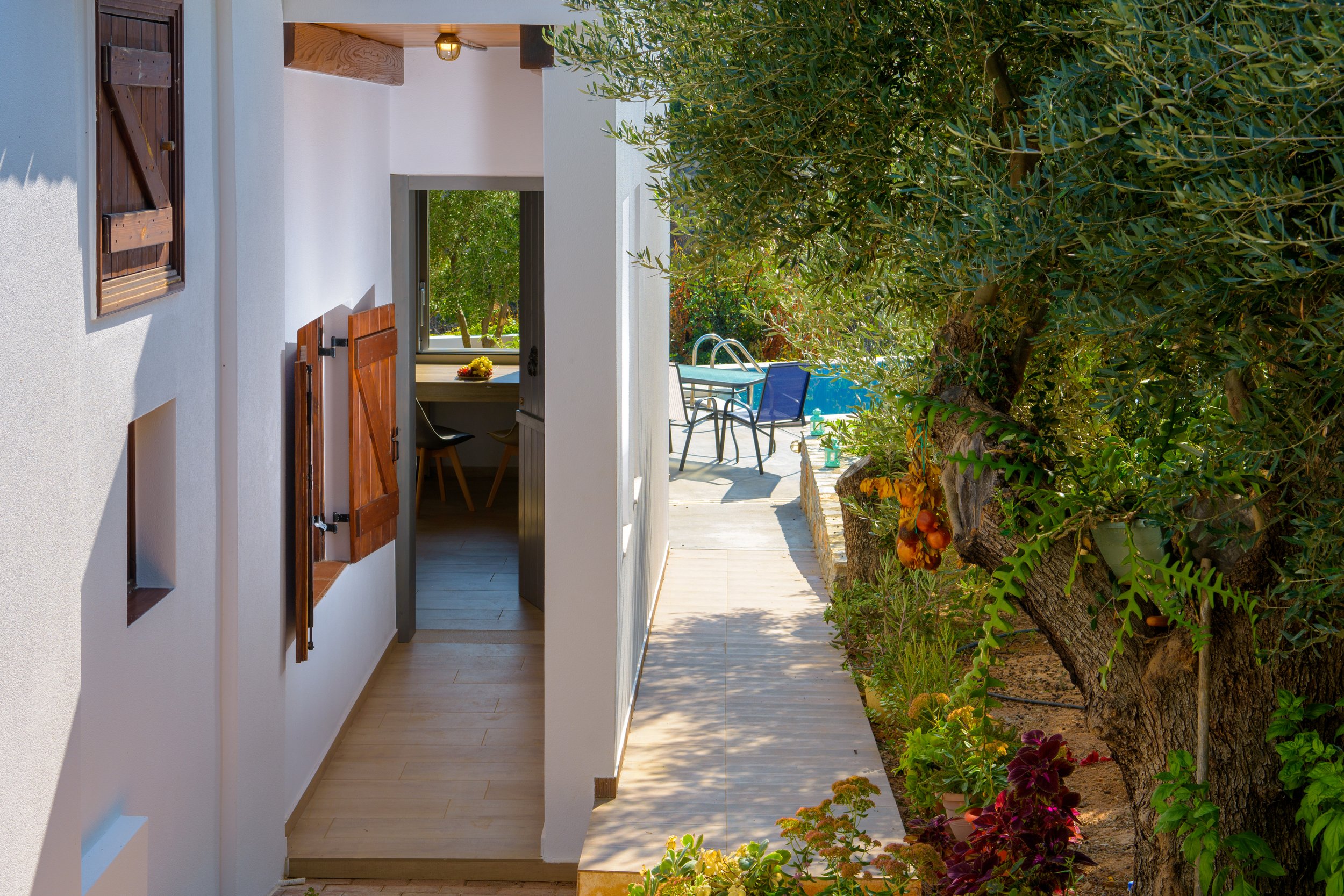 A view of a house entrance hallway with white exterior walls, wooden window shutters, and an open door leading to an outdoor sitting area with a table and chairs. To the right, there is a garden with a large tree and various plants.