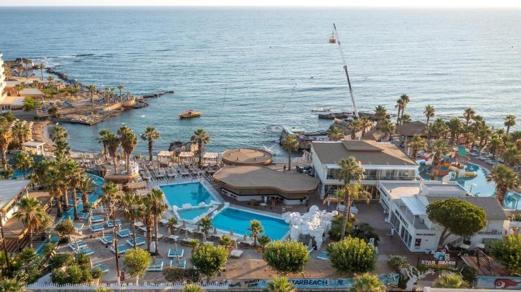 Aerial view of a beachfront resort with pools, palm trees, and beachside amenities near the ocean, with a crane on the dock.
