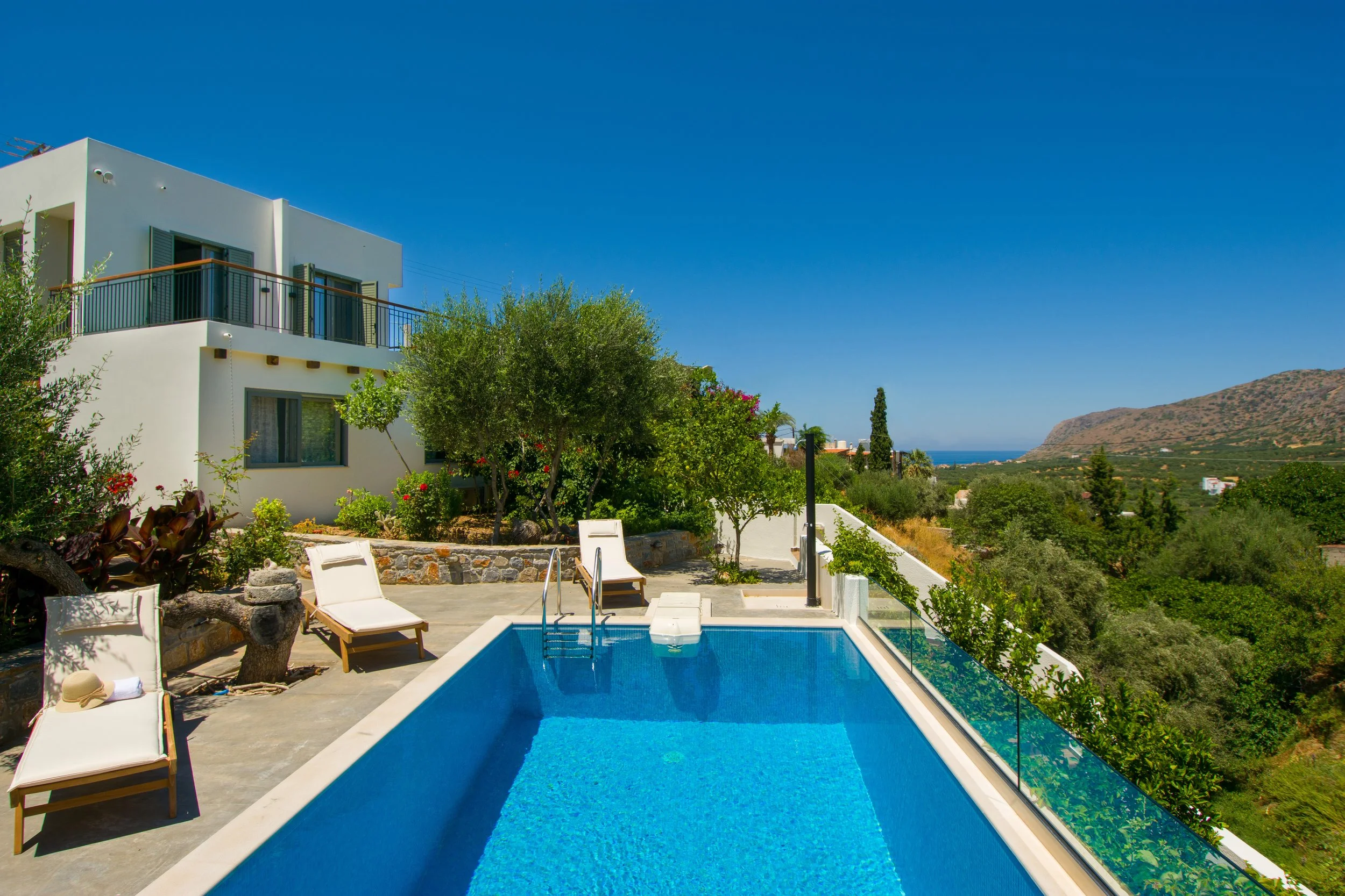 View of a luxurious outdoor swimming pool with lounge chairs, surrounded by greenery, in front of a modern white building with balconies, overlooking hills and ocean in the background under a clear blue sky.