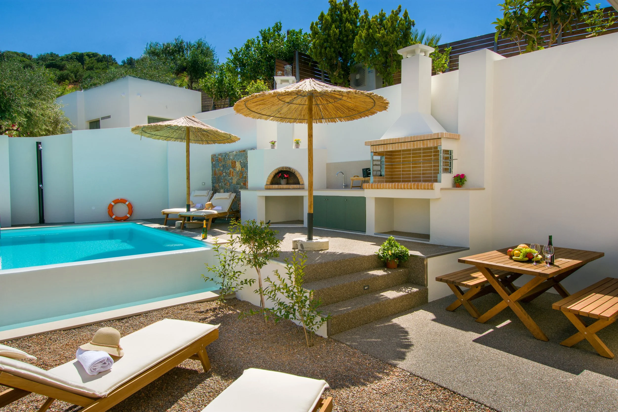 A luxurious outdoor pool area with lounge chairs, straw umbrellas, and a small outdoor fireplace, surrounded by white walls and lush greenery, under a clear blue sky.