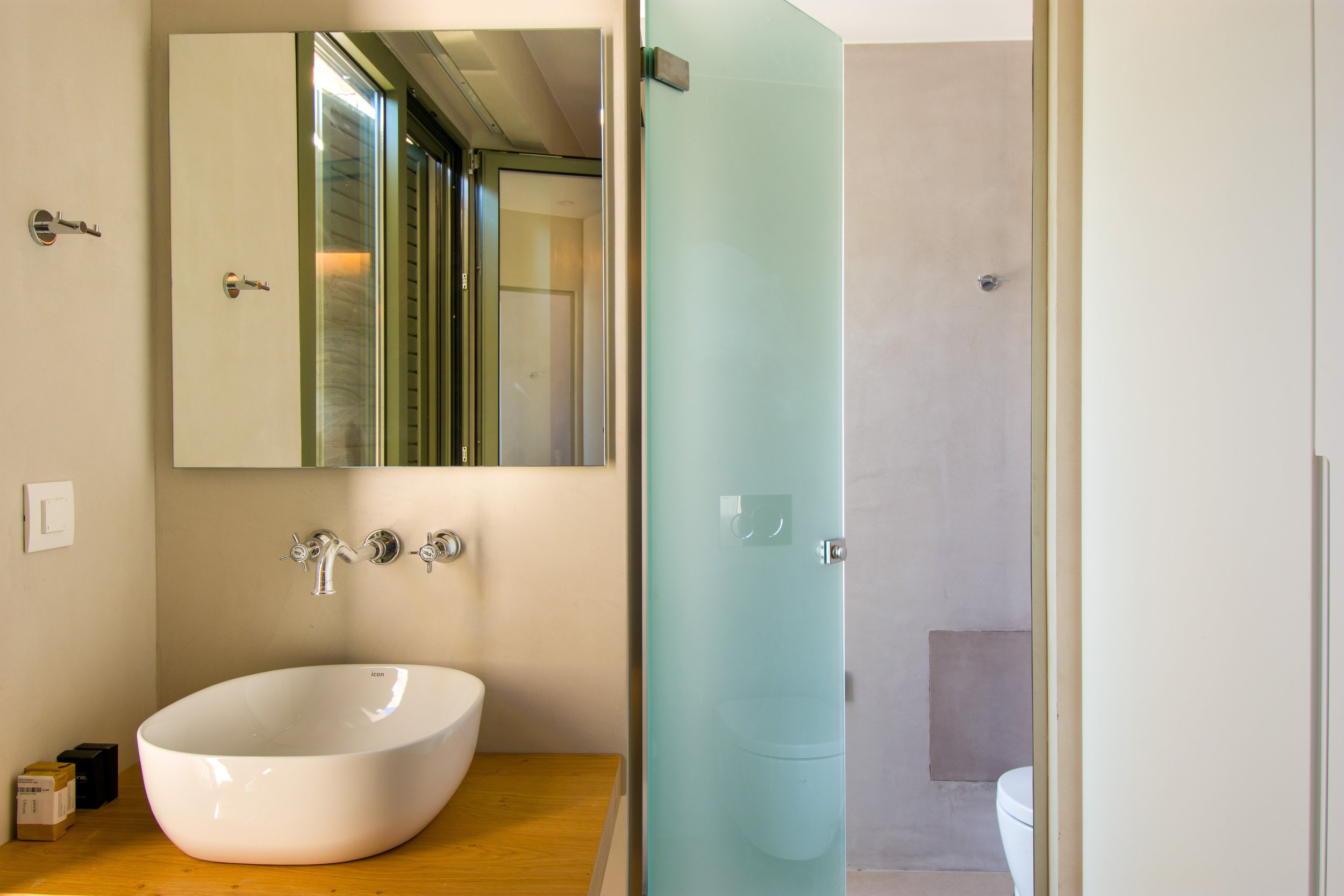 Modern bathroom with a white oval vessel sink on a wooden countertop, a large mirror, and a light-colored tiled shower with a glass door.