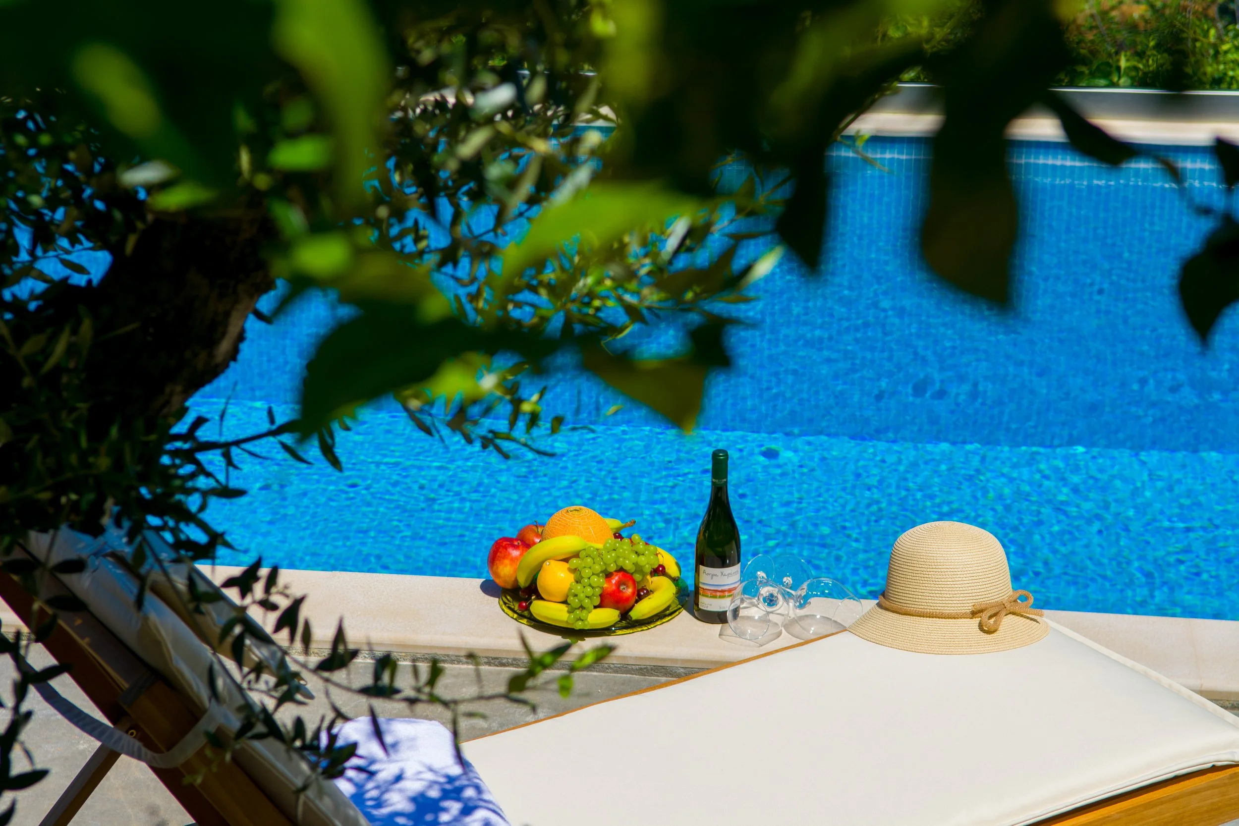 A table set with fresh fruit, wine, and a sun hat, by a swimming pool surrounded by greenery.