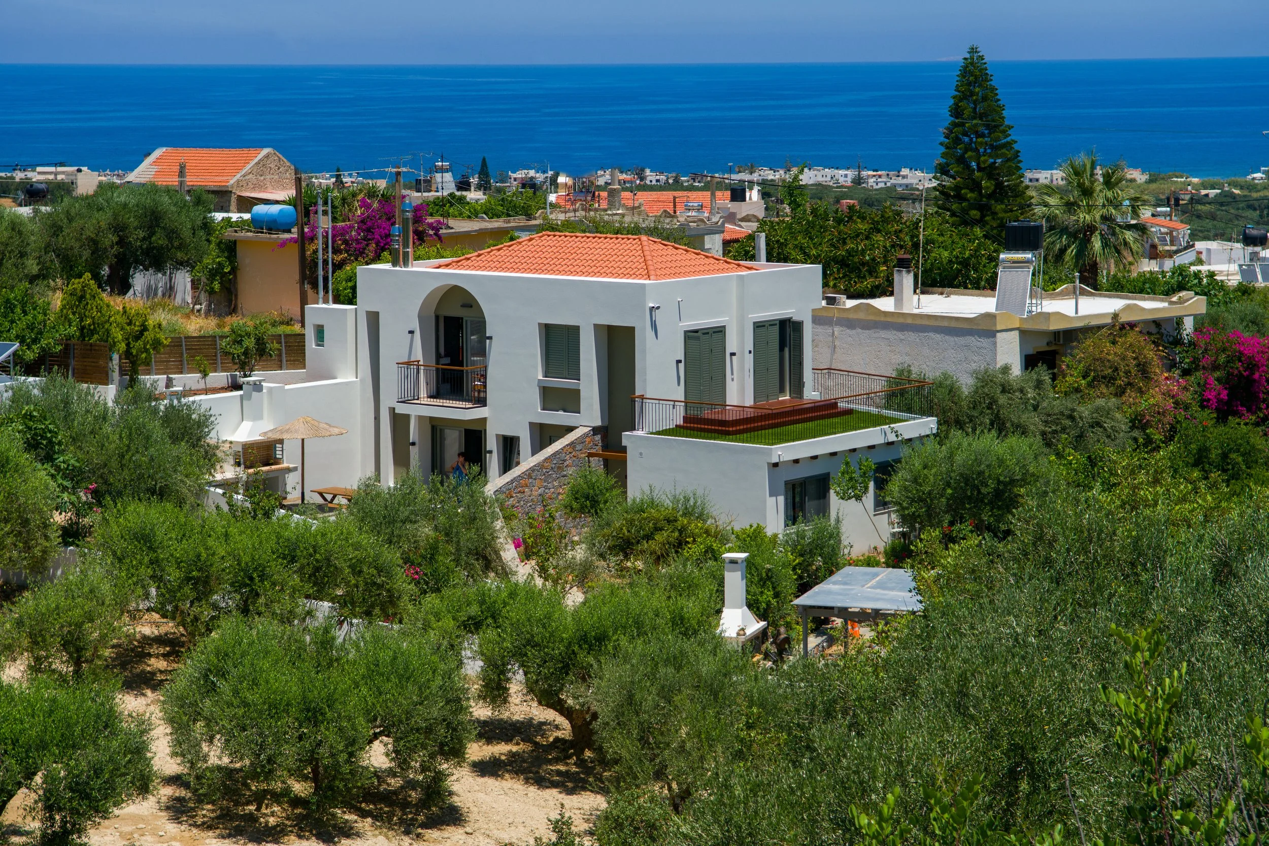 A residential area near the coast with white modern houses, orange-tiled roofs, and lush greenery, overlooking the blue sea in the background.