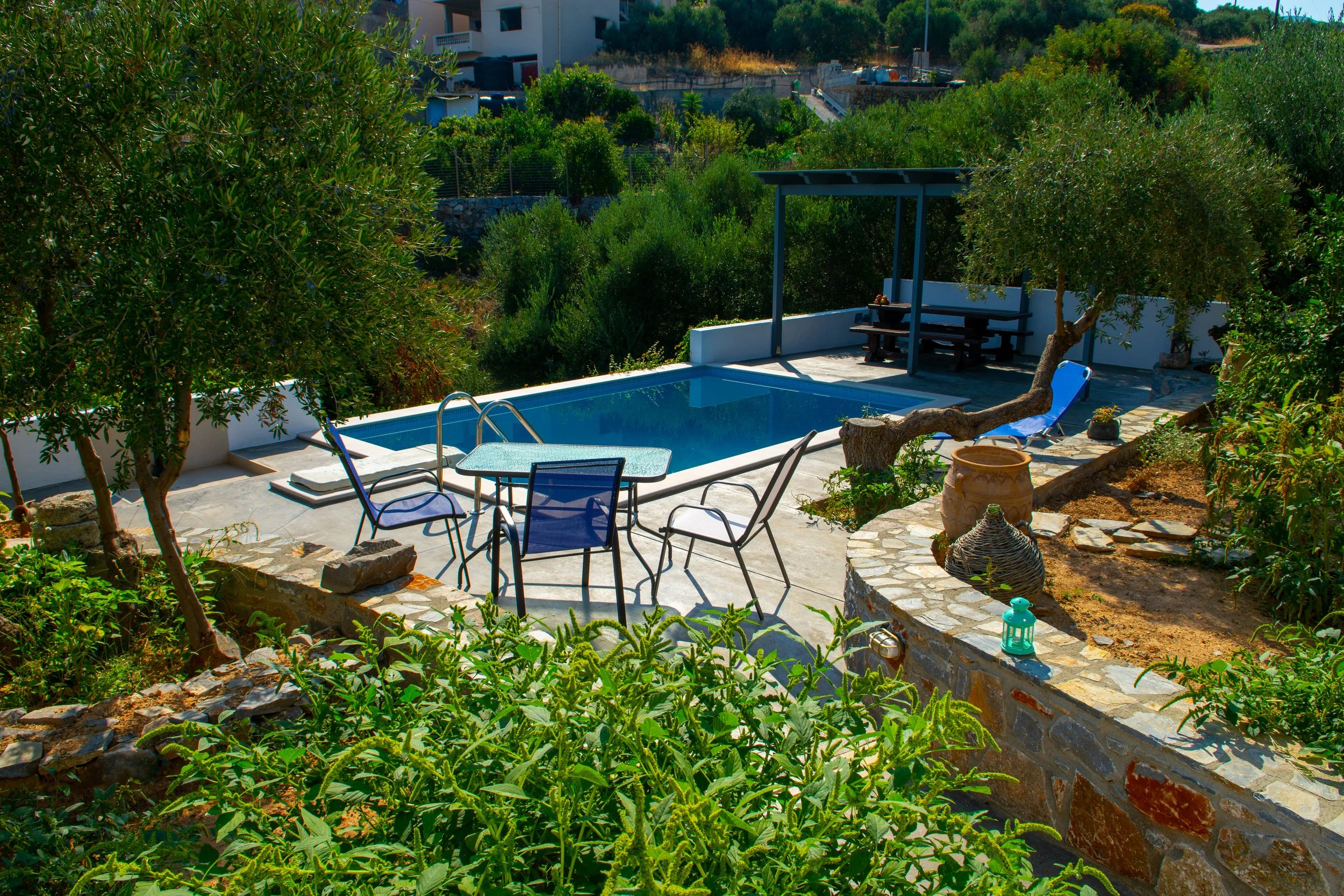 A backyard scene featuring a small swimming pool with a ladder, surrounded by outdoor furniture including chairs and a table, lush greenery with various trees and plants, and a shaded seating area with a bench and table. There are also arrangements o