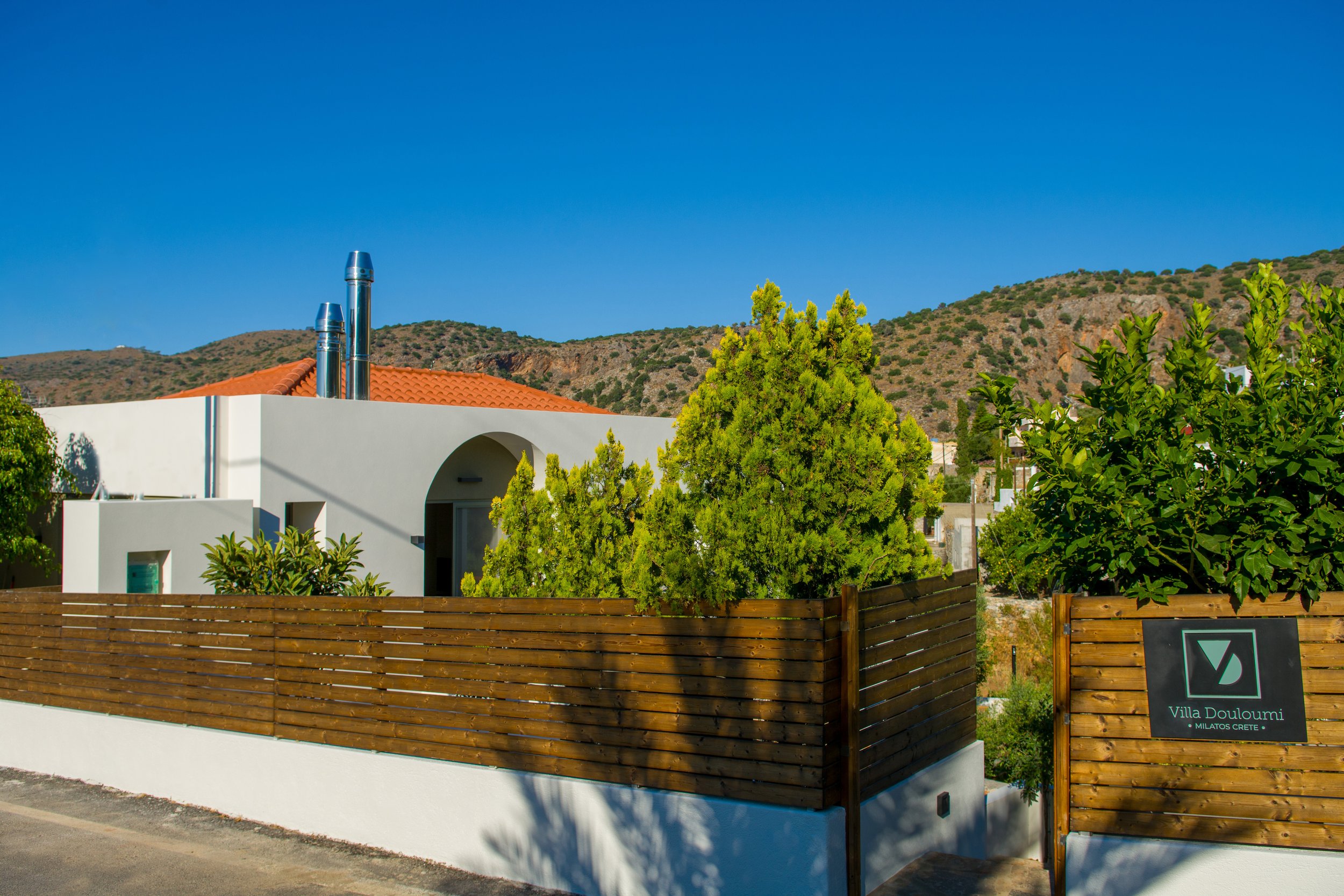 Modern white villa with a red-tiled roof, surrounded by green trees, and a wooden fence in front; mountains in the background under a clear blue sky.