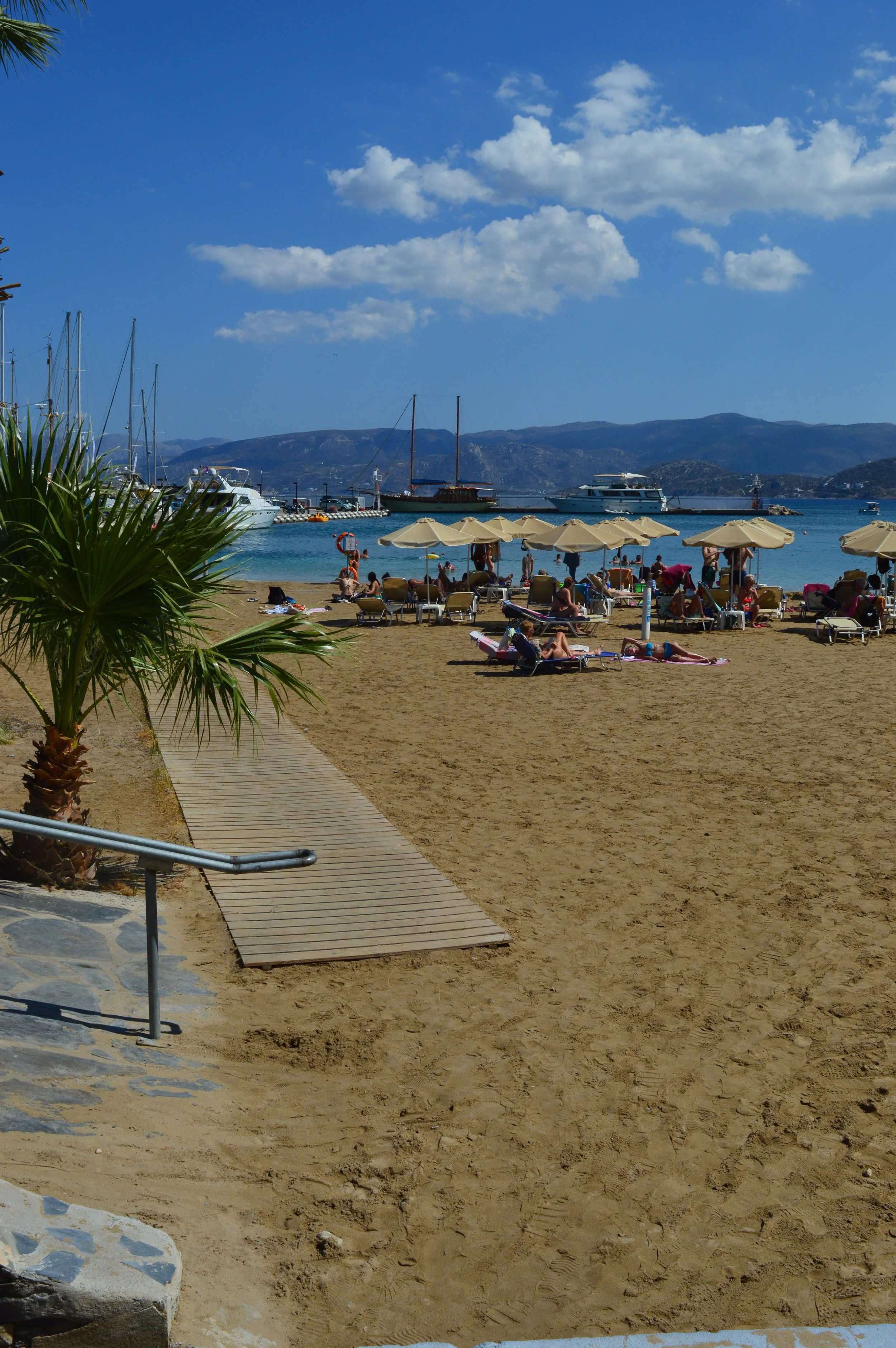 Beach scene with people under umbrellas, boats docked in the water, sandy shore, mountains in the background, partly cloudy sky.