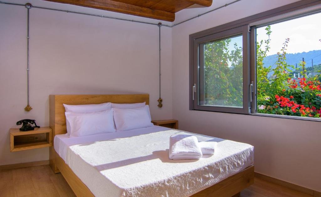 A minimalistic bedroom with a bed, white linens, and two neatly folded towels on top. There are two pillows, a wooden headboard, and a small wooden nightstand with a vintage black rotary phone. The room has white walls, a large window with a view of 