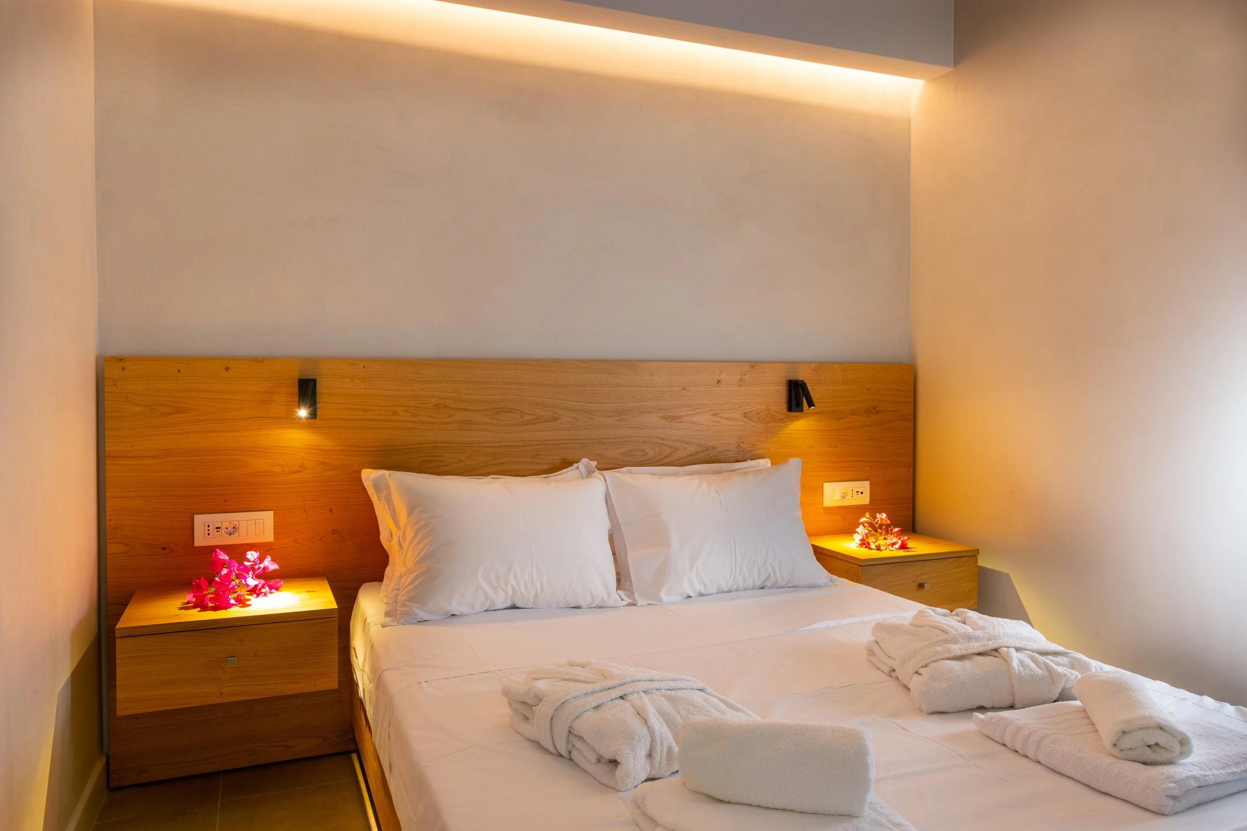 A neatly made hotel bed with white linens, two pillows, and towels shaped like swans. There are two small bedside tables with pink flowers and a lit lamp on each. The headboard is wooden, and there are black reading lights attached to it. The wall ab