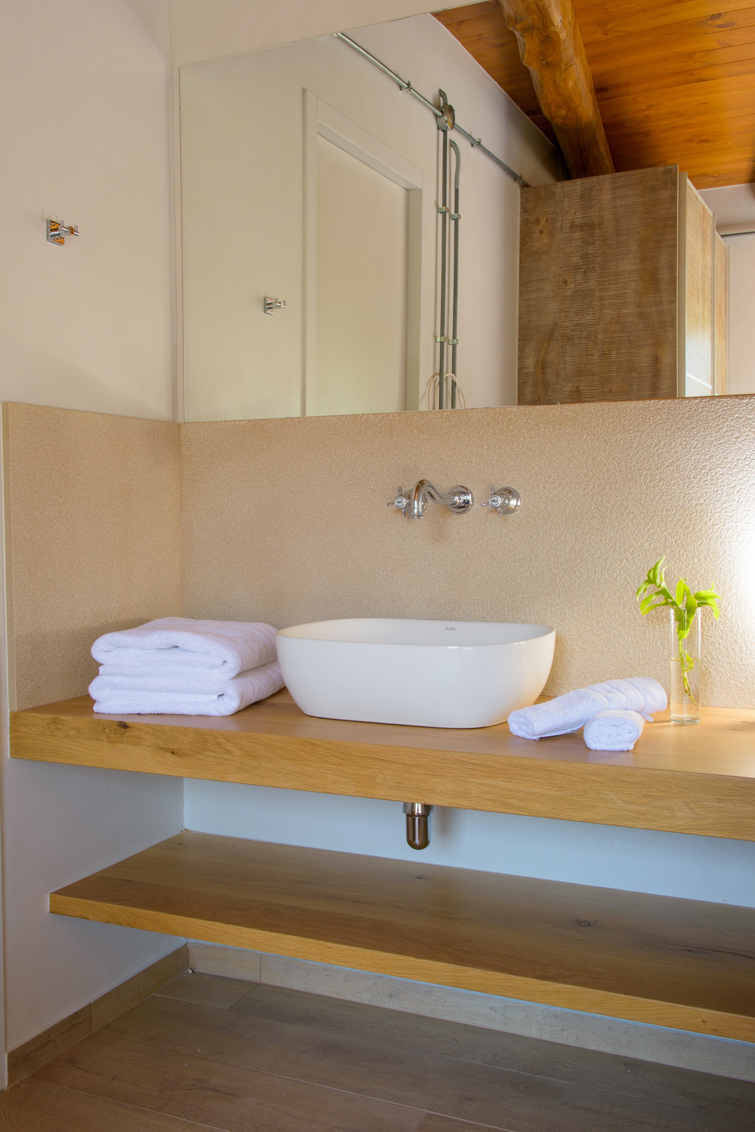 A bathroom with a wooden countertop, a white vessel sink, and towels. There is a small plant in a glass next to the sink and a mirror behind it. The wall behind the countertop is beige, with a textured finish.