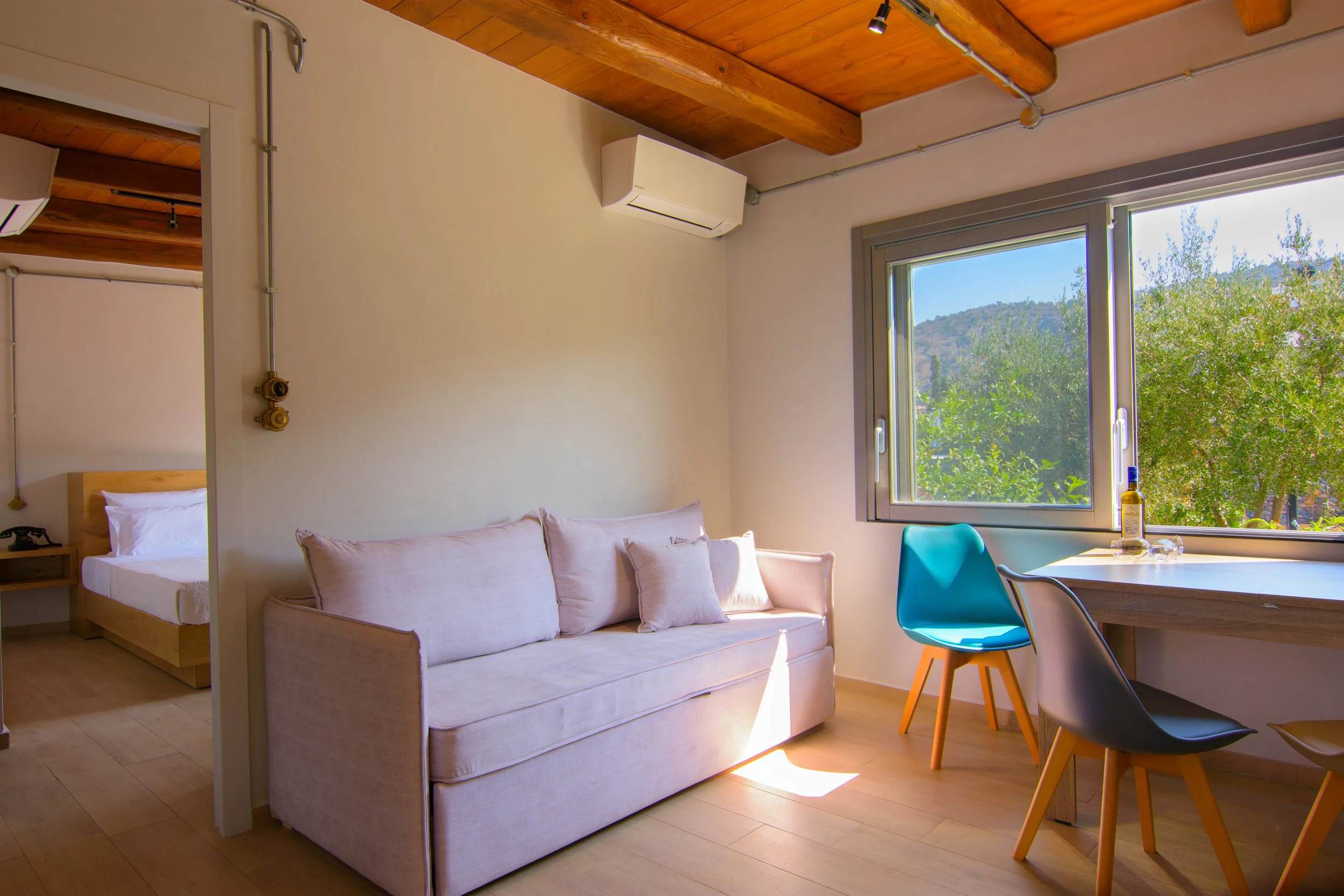 Modern apartment interior with a beige sofa and colorful chairs near a large window showing outdoor trees and hills.