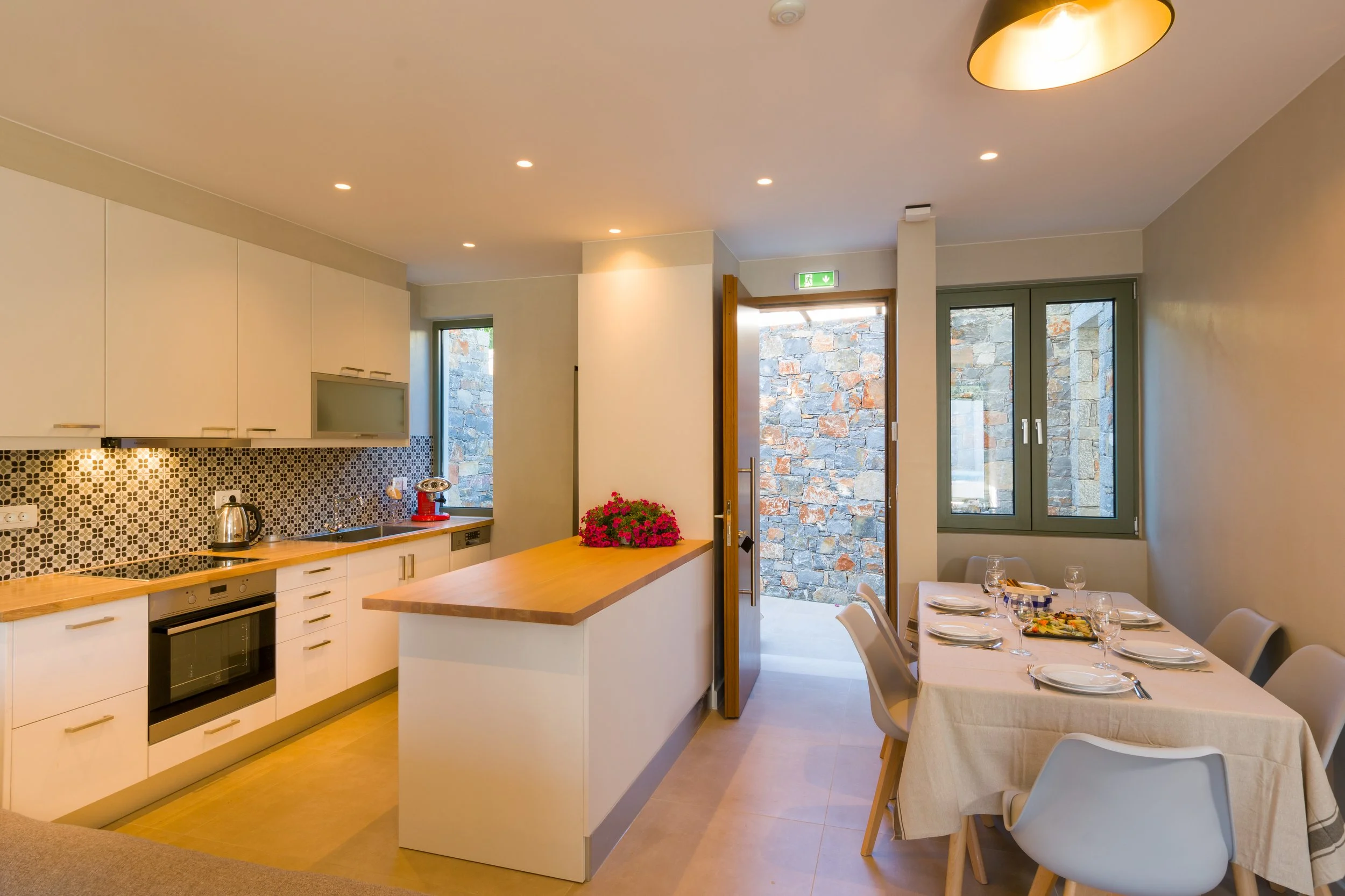 A modern kitchen and dining area with white cabinets, a patterned backsplash, a wooden countertop, and a table set for dinner with white chairs and tableware. Large windows and a door lead to an outdoor stone wall.