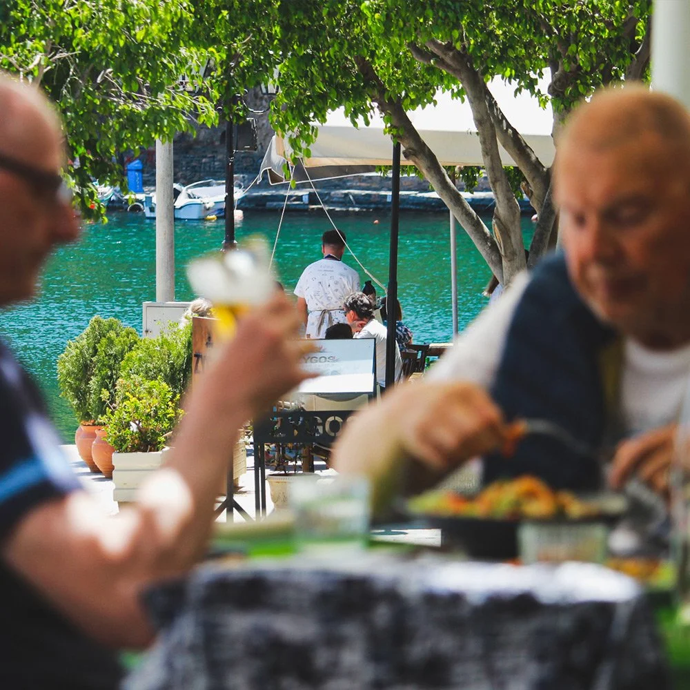 People dining at an outdoor restaurant near a waterfront with boats, green water, trees, and a blue sky in the background.