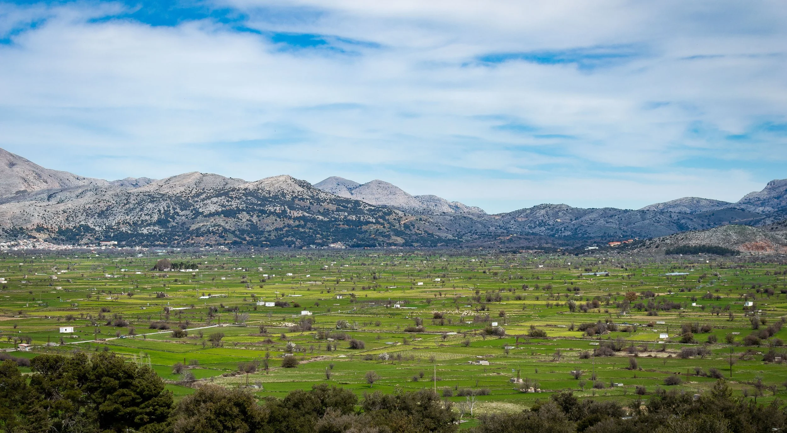 Panoramic view of a rural valley with green fields and scattered trees, backed by mountains under a partly cloudy sky.