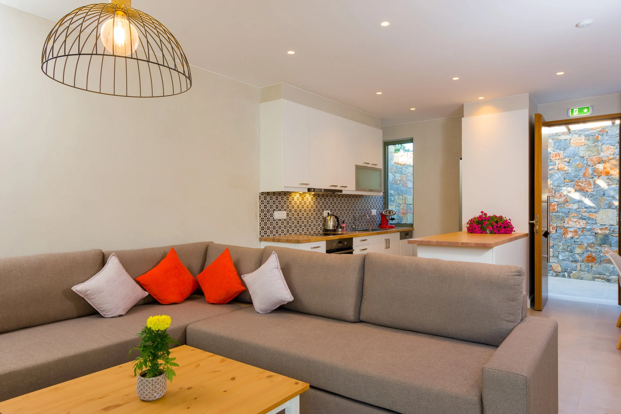 Modern open-concept living room with a beige sectional sofa topped with orange and pink pillows, a wooden coffee table with a potted yellow flowering plant, a kitchen with white cabinets, a patterned backsplash, and a granite countertop, and a stone 